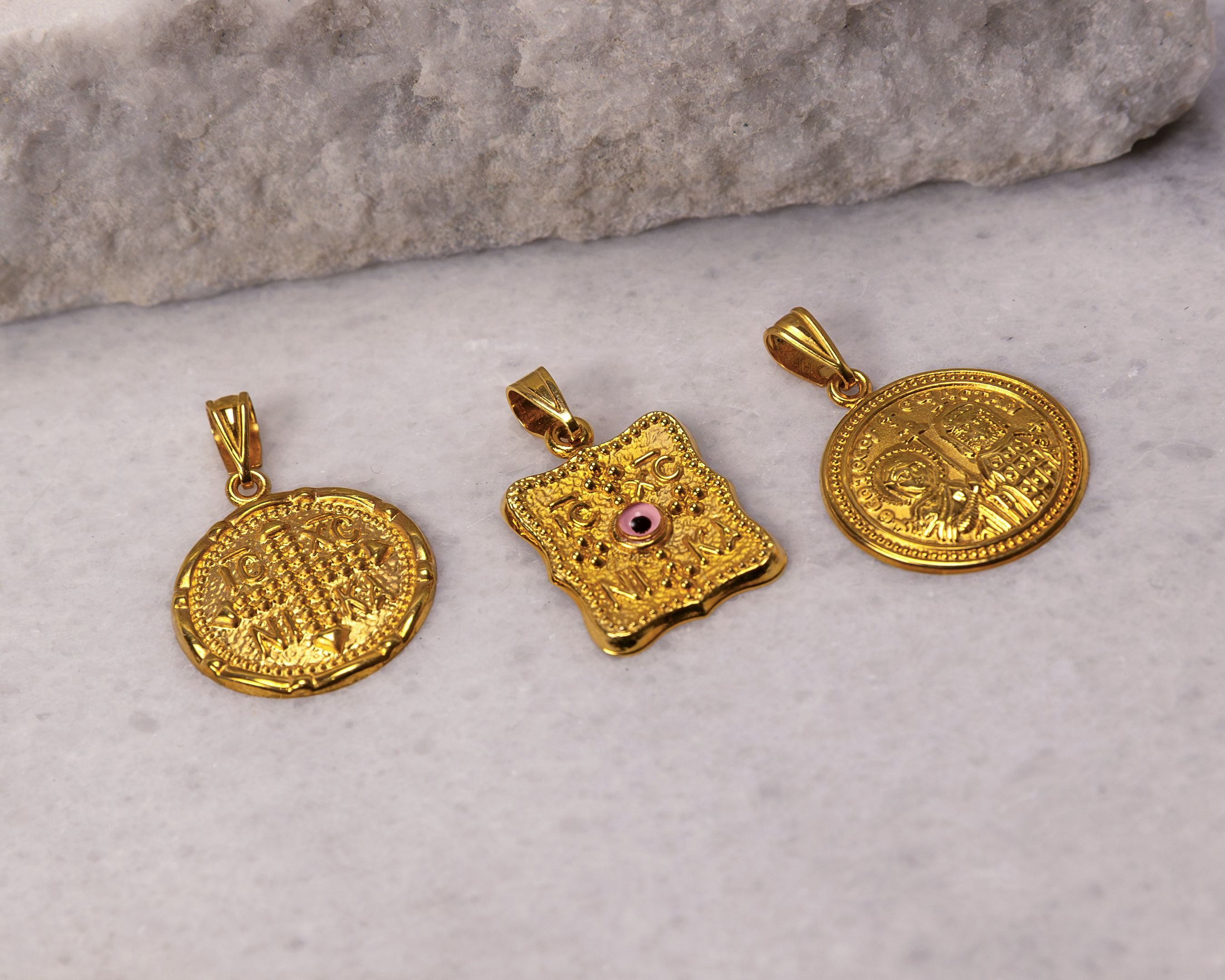Three gold religious pendants with intricate designs placed on a light marble surface near a stone background.