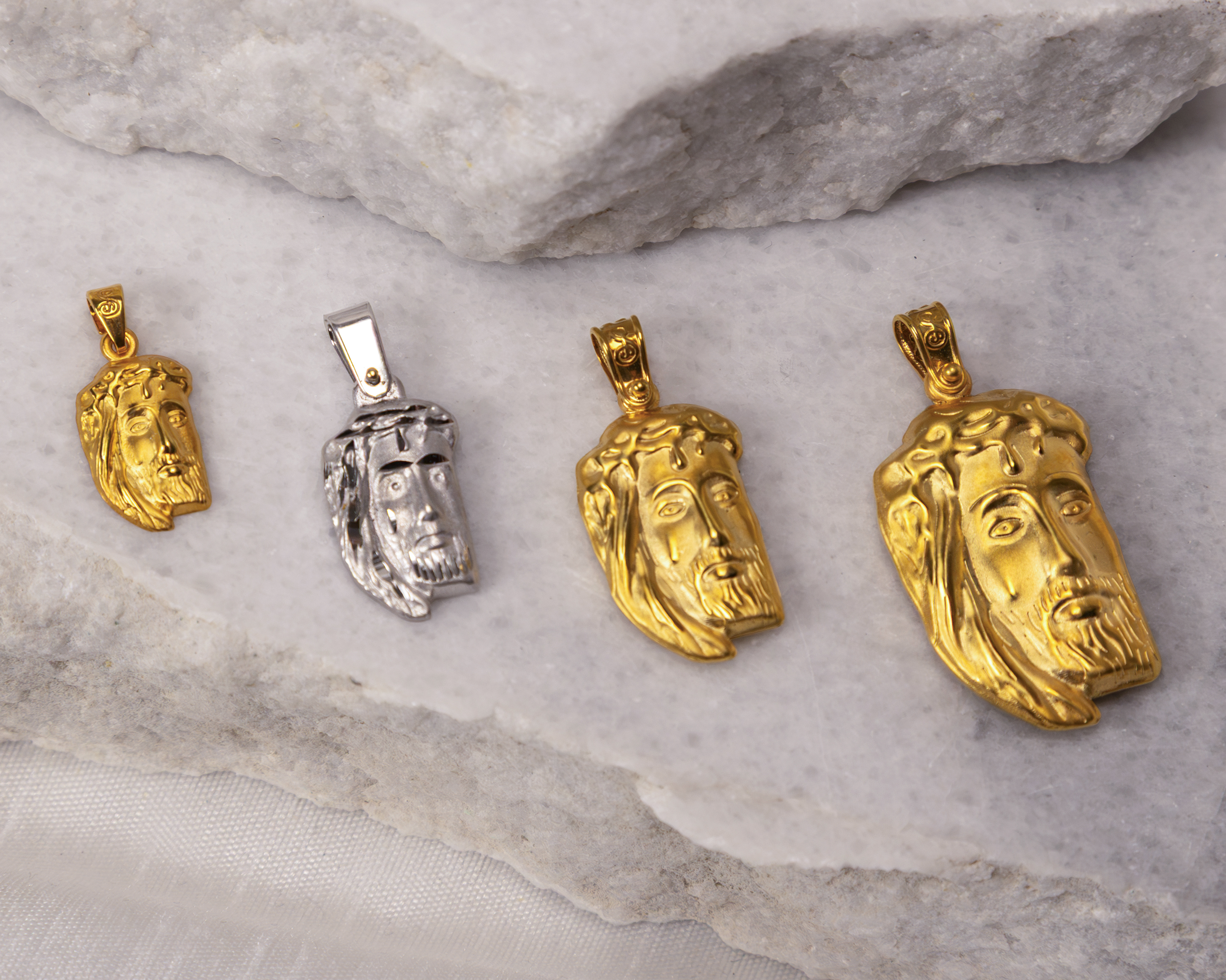 Four Jesus head pendants in varying sizes and finishes, three gold and one silver, arranged on a light stone surface.