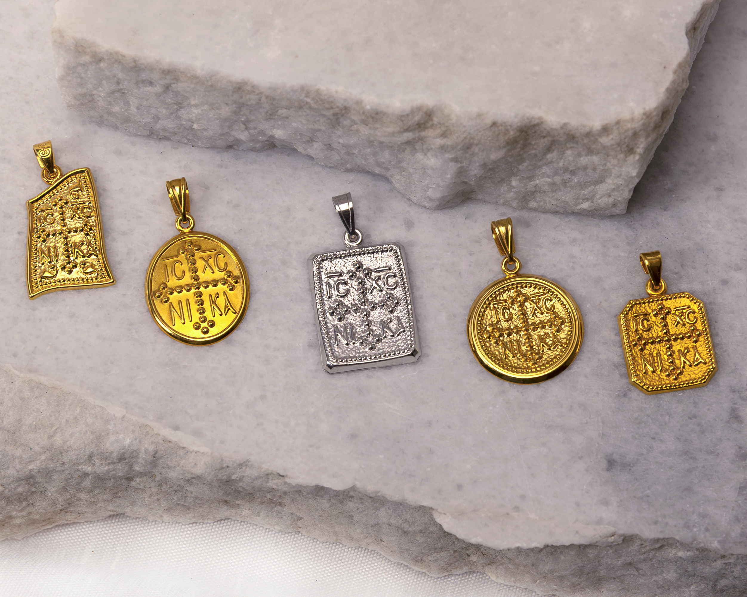 Five gold and silver religious pendants with IC XC NIKA inscriptions on a textured stone surface.