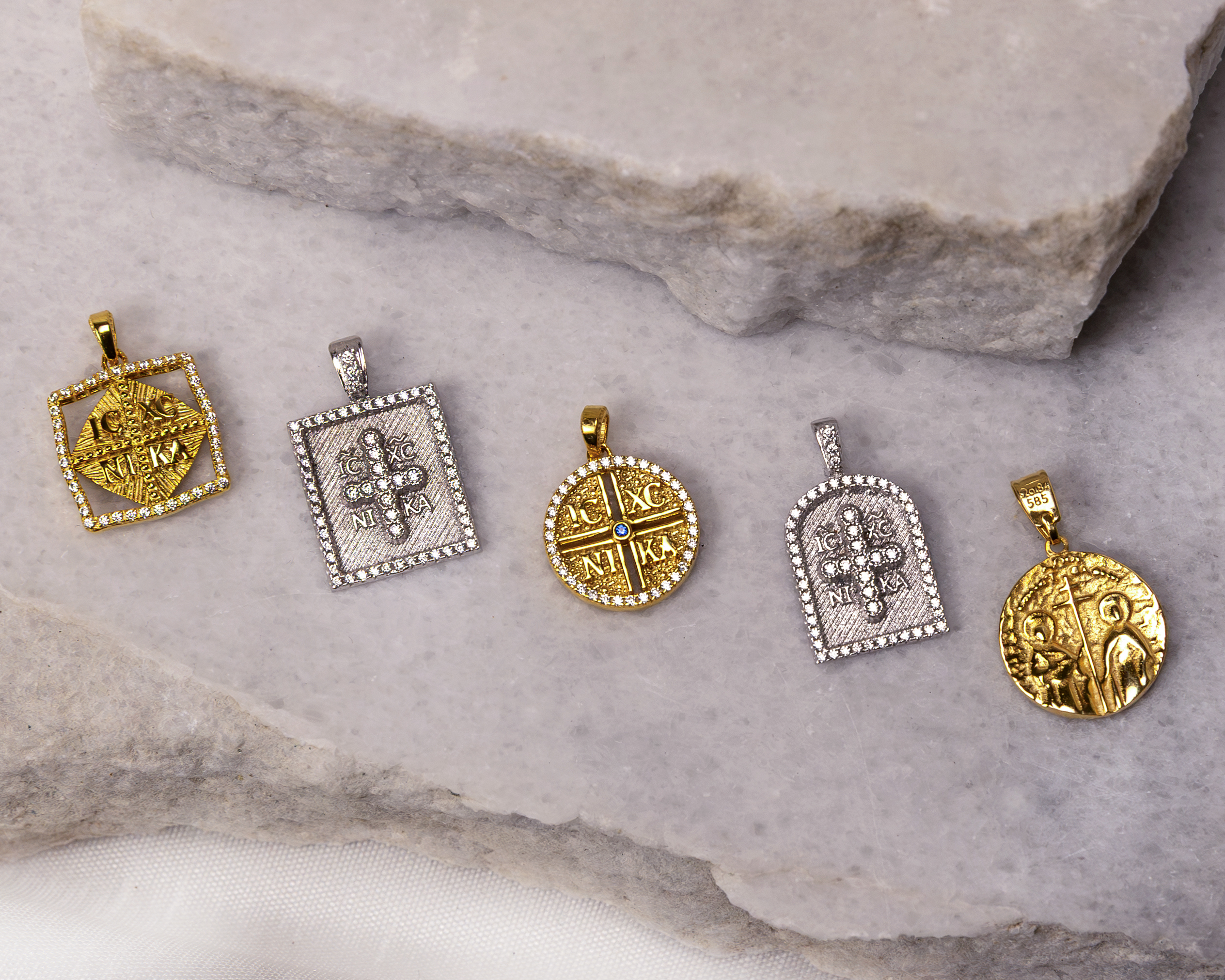 Five gold and silver religious pendants with cross designs and inscriptions displayed on a light gray stone surface.