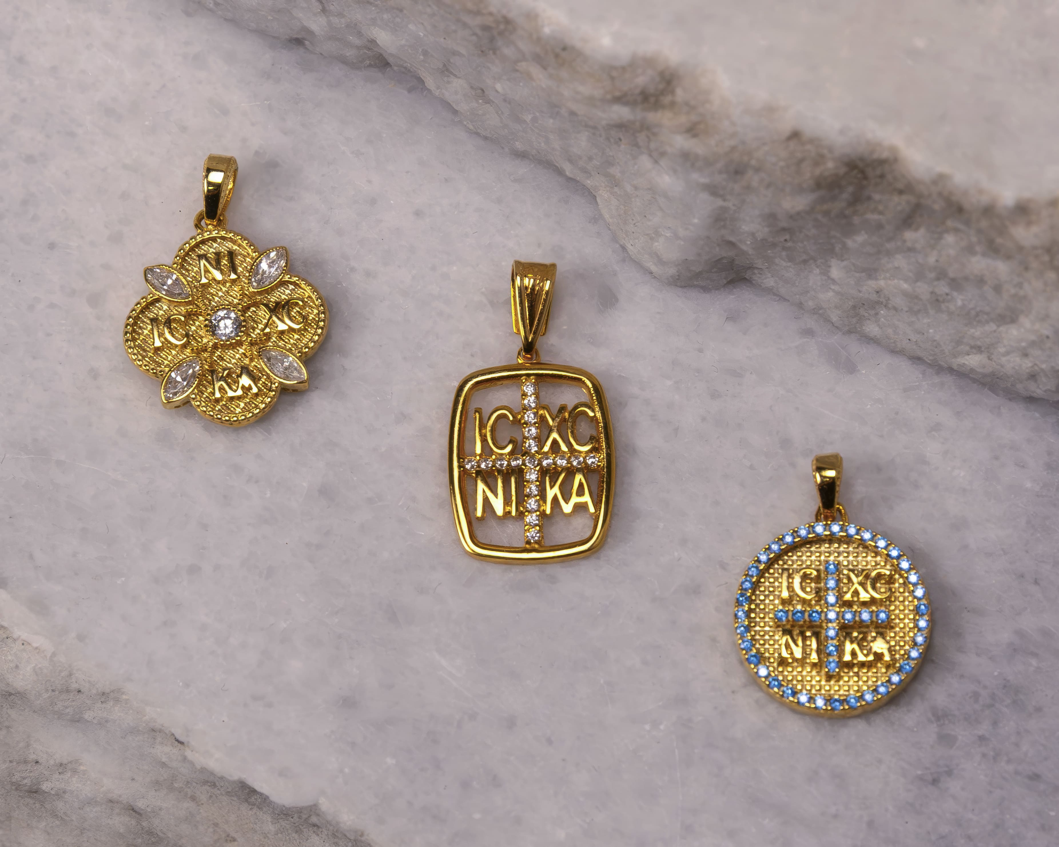 Three gold religious pendants with IC XC NI KA inscriptions and decorated with clear and blue gemstones, placed on a marble surface.