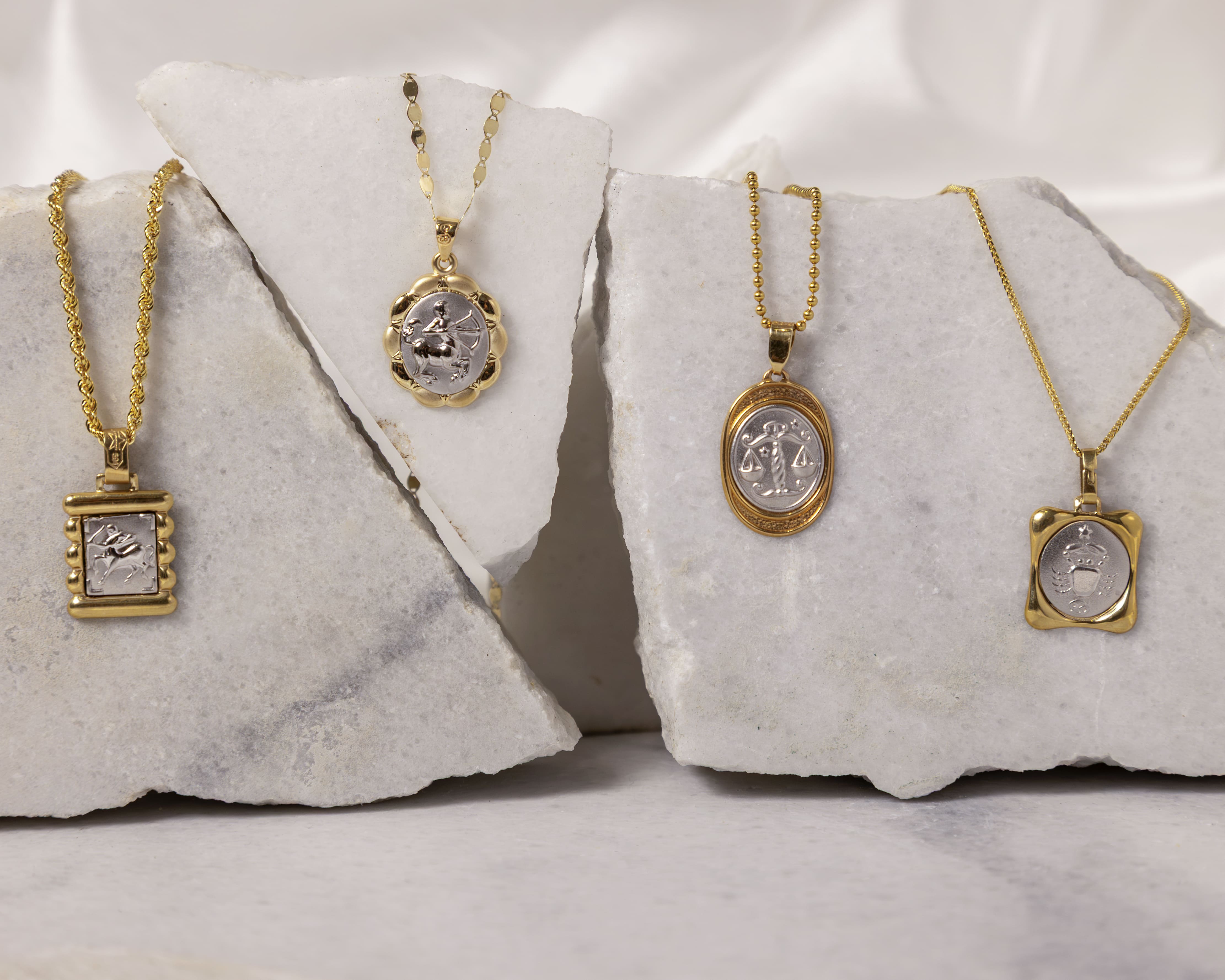 Four gold and silver zodiac sign pendants displayed on white marble slabs.