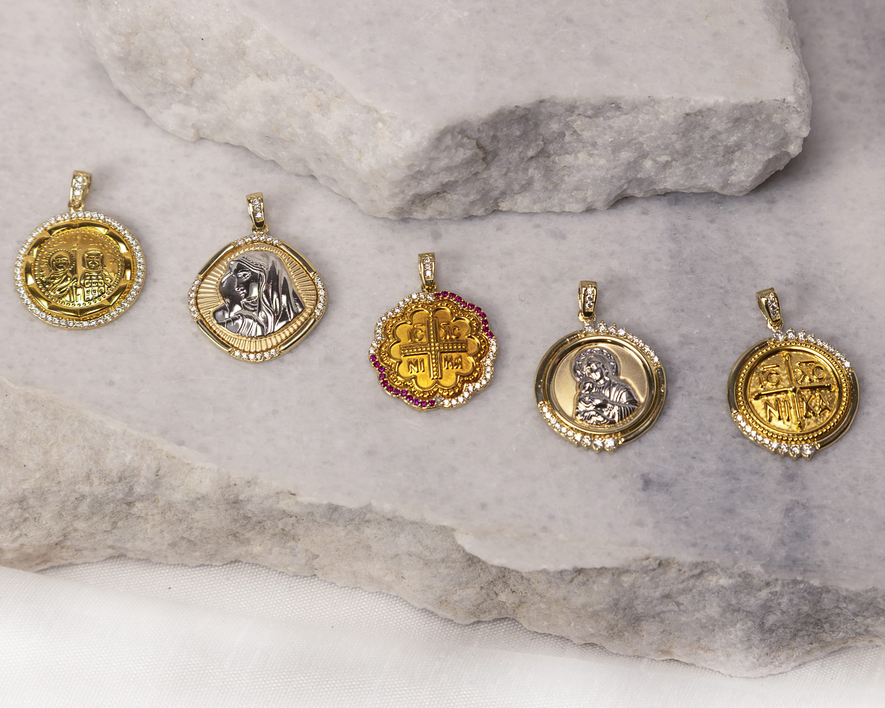 Five gold and silver religious pendants with crystal embellishments on a marble surface.