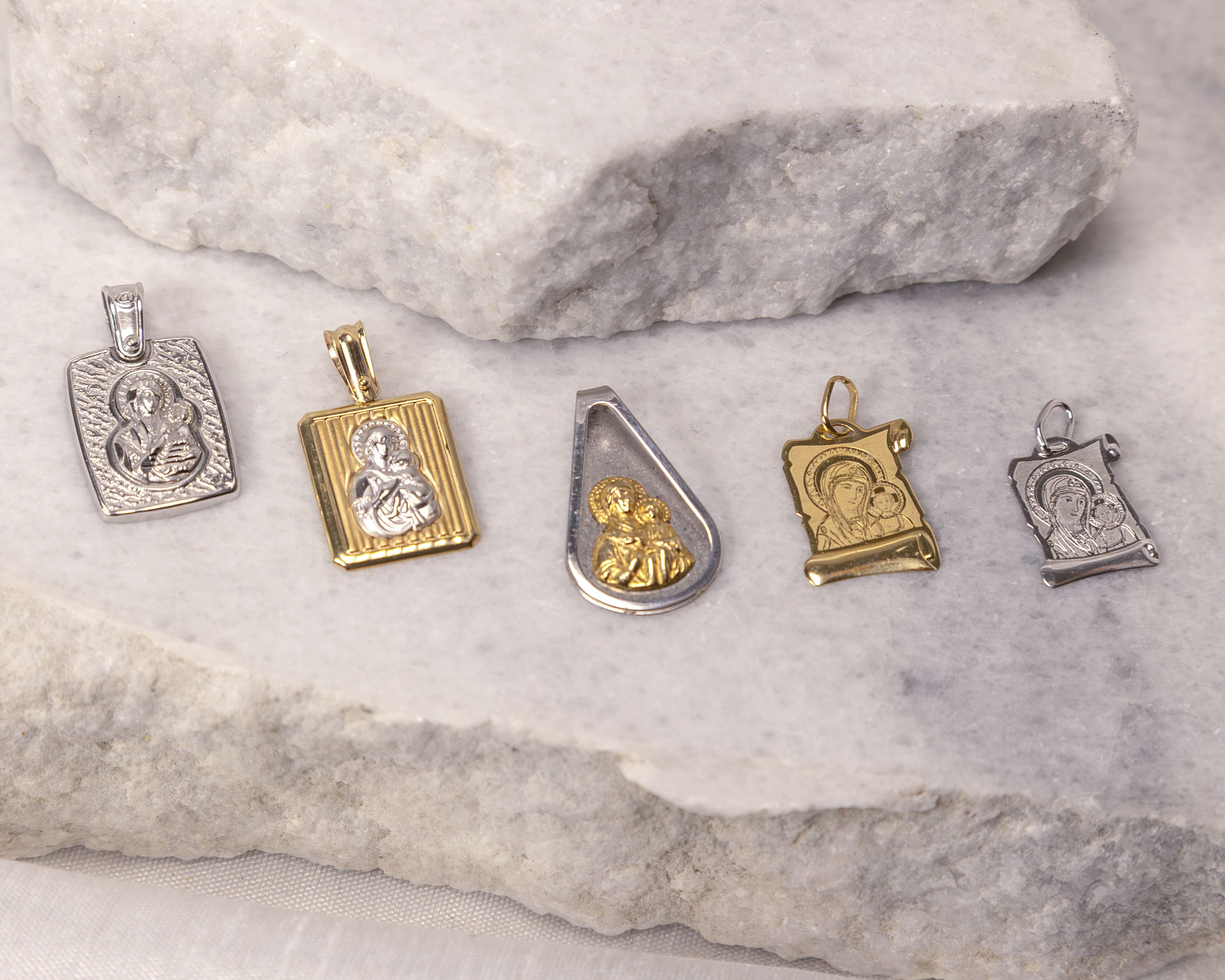 Five religious pendants depicting the Virgin Mary and child, in silver and gold, arranged on a white marble surface.