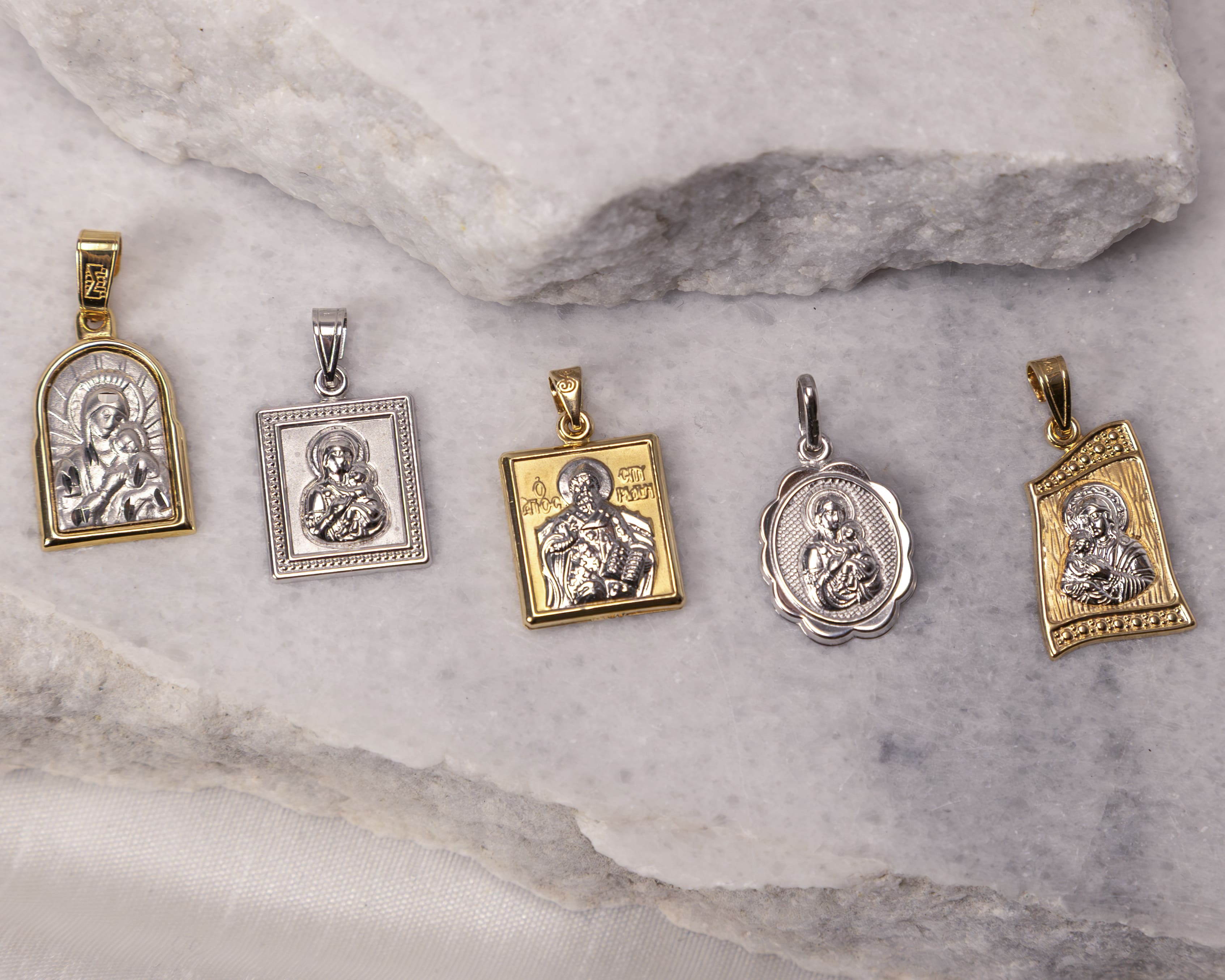 Five religious gold and silver pendants featuring detailed Christian iconography, placed on white marble slabs.