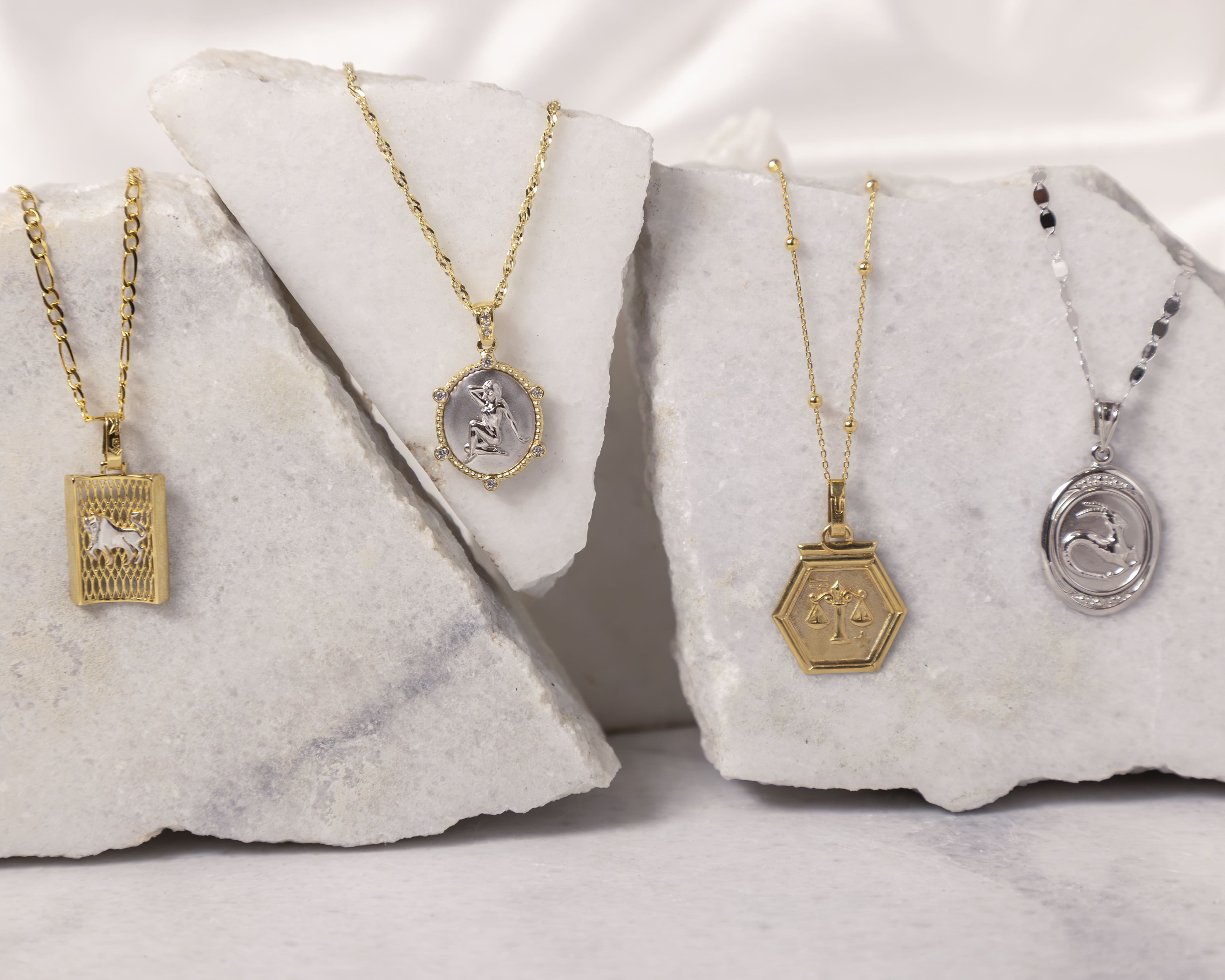 Four gold and silver zodiac sign necklaces displayed on white marble pieces, featuring Aries, Virgo, Libra, and Capricorn signs.