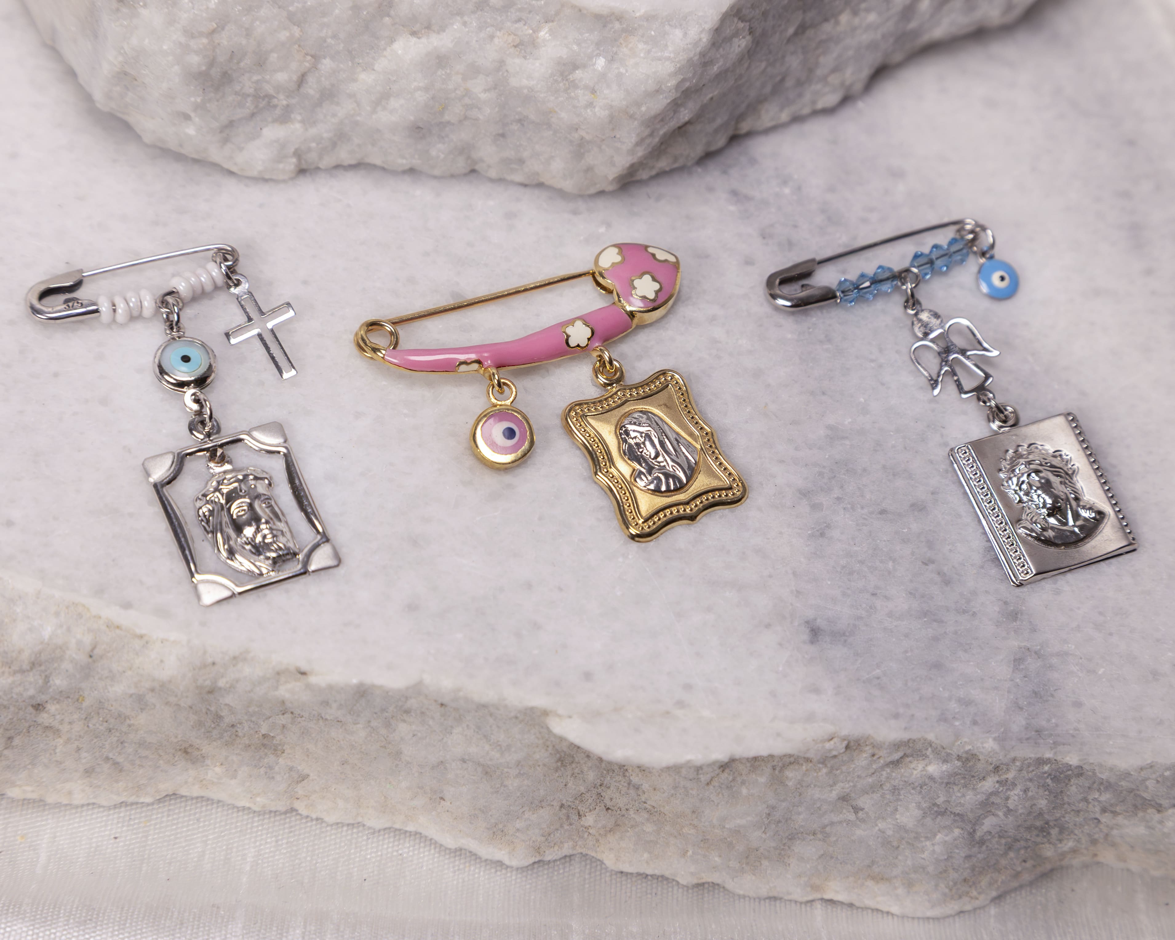 Three decorative pins with religious charms including crosses, blue eye beads, and engraved religious figures on a marble surface.