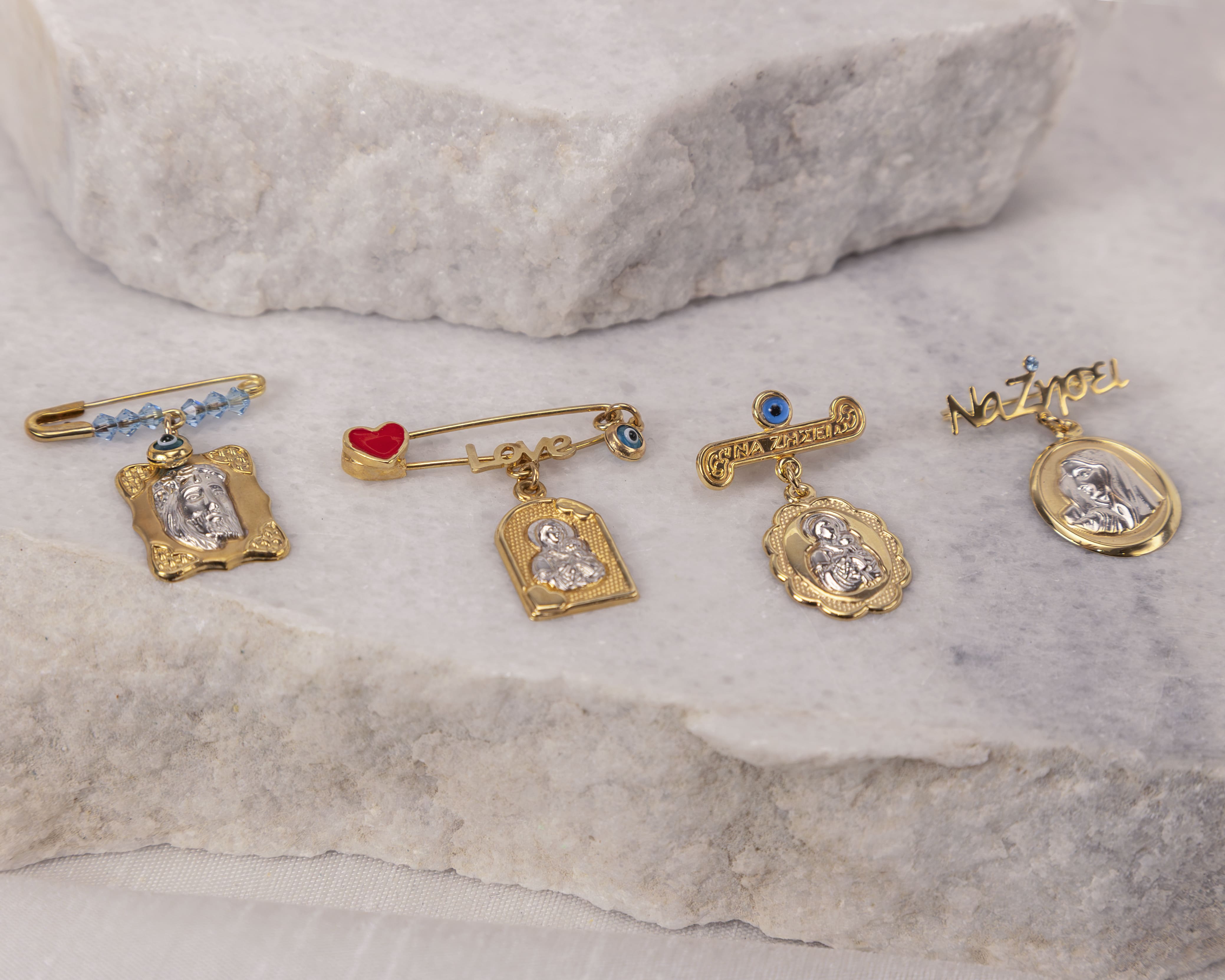 Four gold safety pin brooches with religious charms and blue evil eye beads on a light marble surface.
