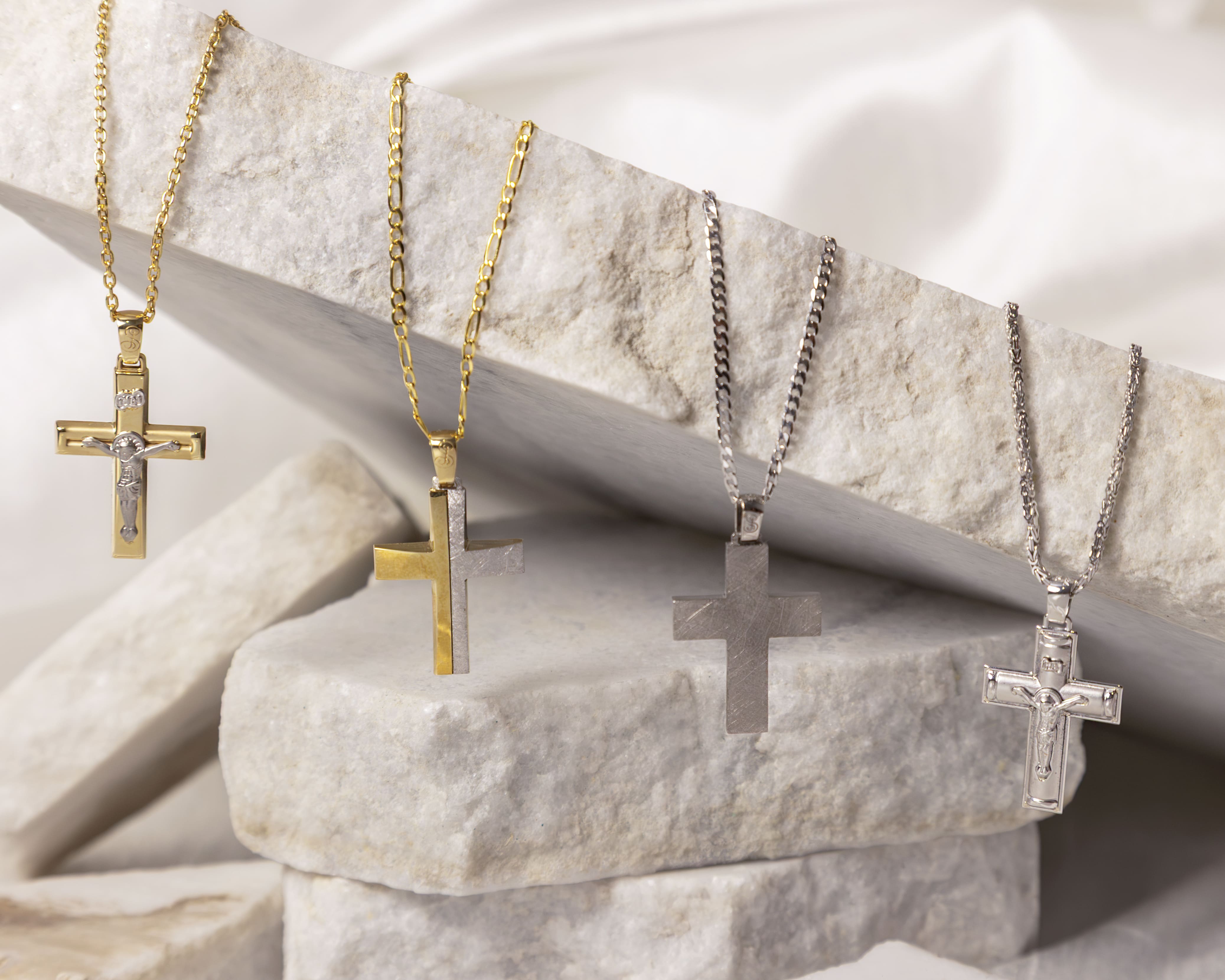 Four cross necklaces with gold and silver chains displayed on textured stone blocks.