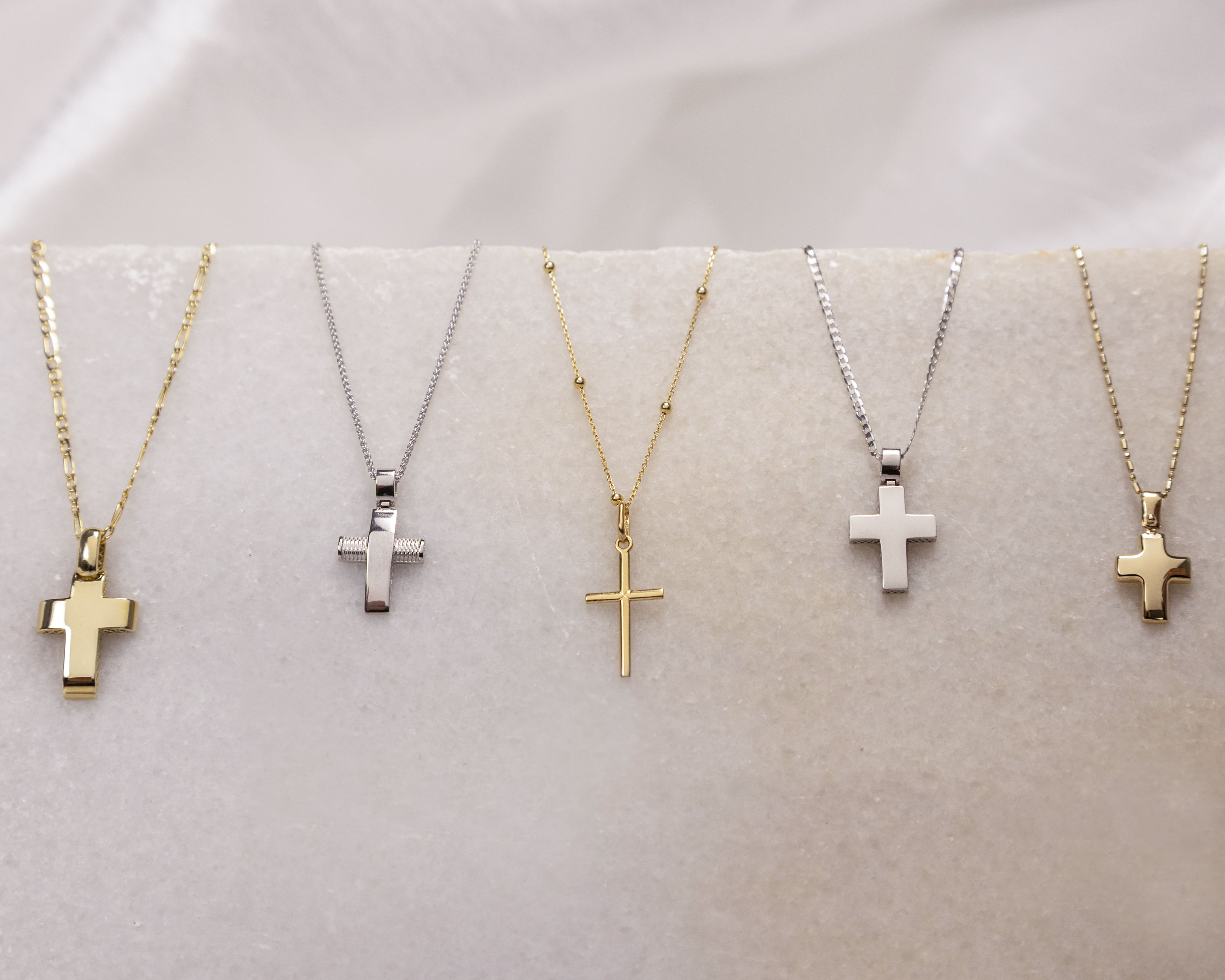 Five cross necklaces on delicate gold and silver chains displayed on a light marble surface.