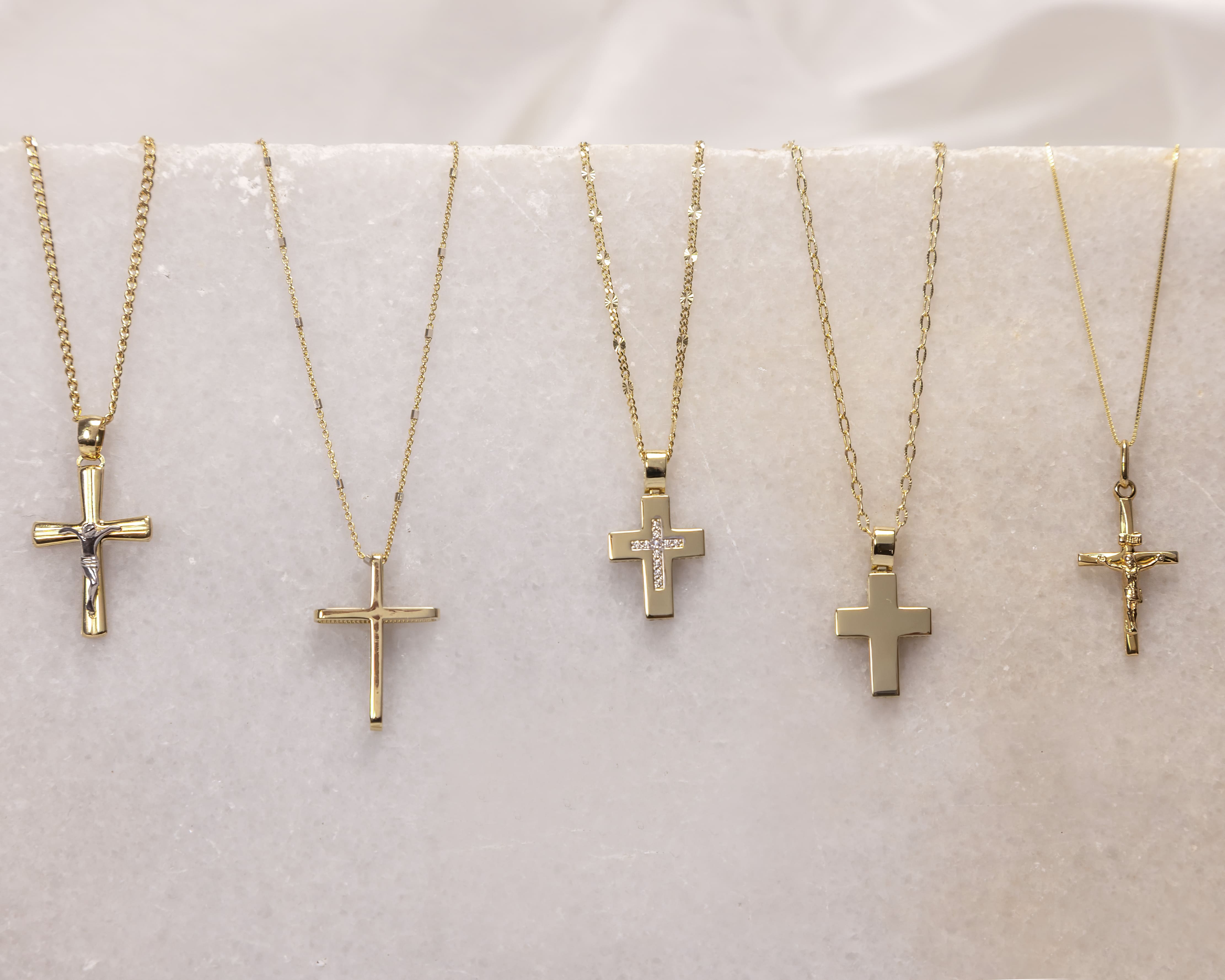 Five gold cross necklaces with different designs displayed on a white marble surface.