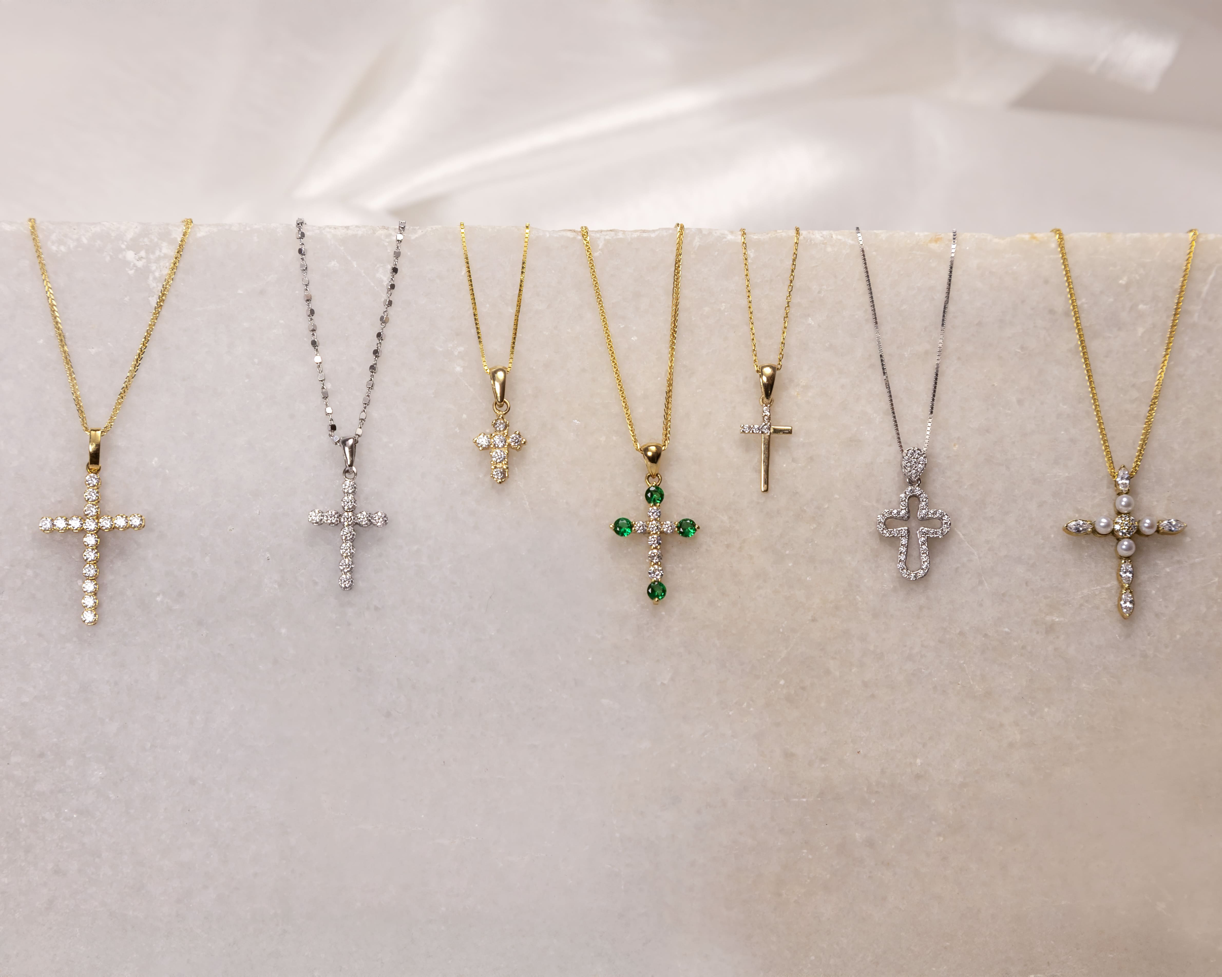 Seven cross pendant necklaces displayed on a light marble surface, featuring various designs with gold and silver chains, some adorned with diamonds, green gems, and pearls.