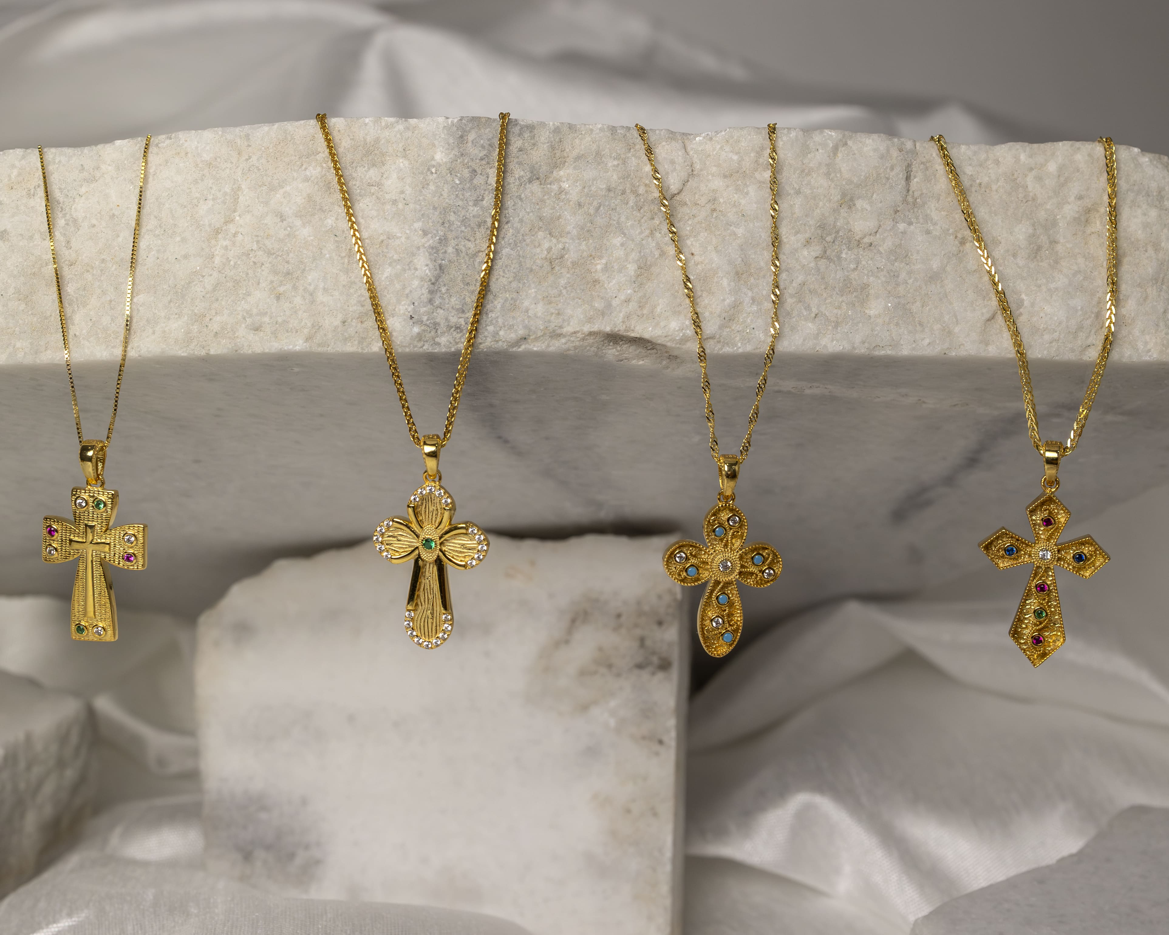 Four gold cross necklaces with colorful gemstone accents displayed on a textured stone slab.