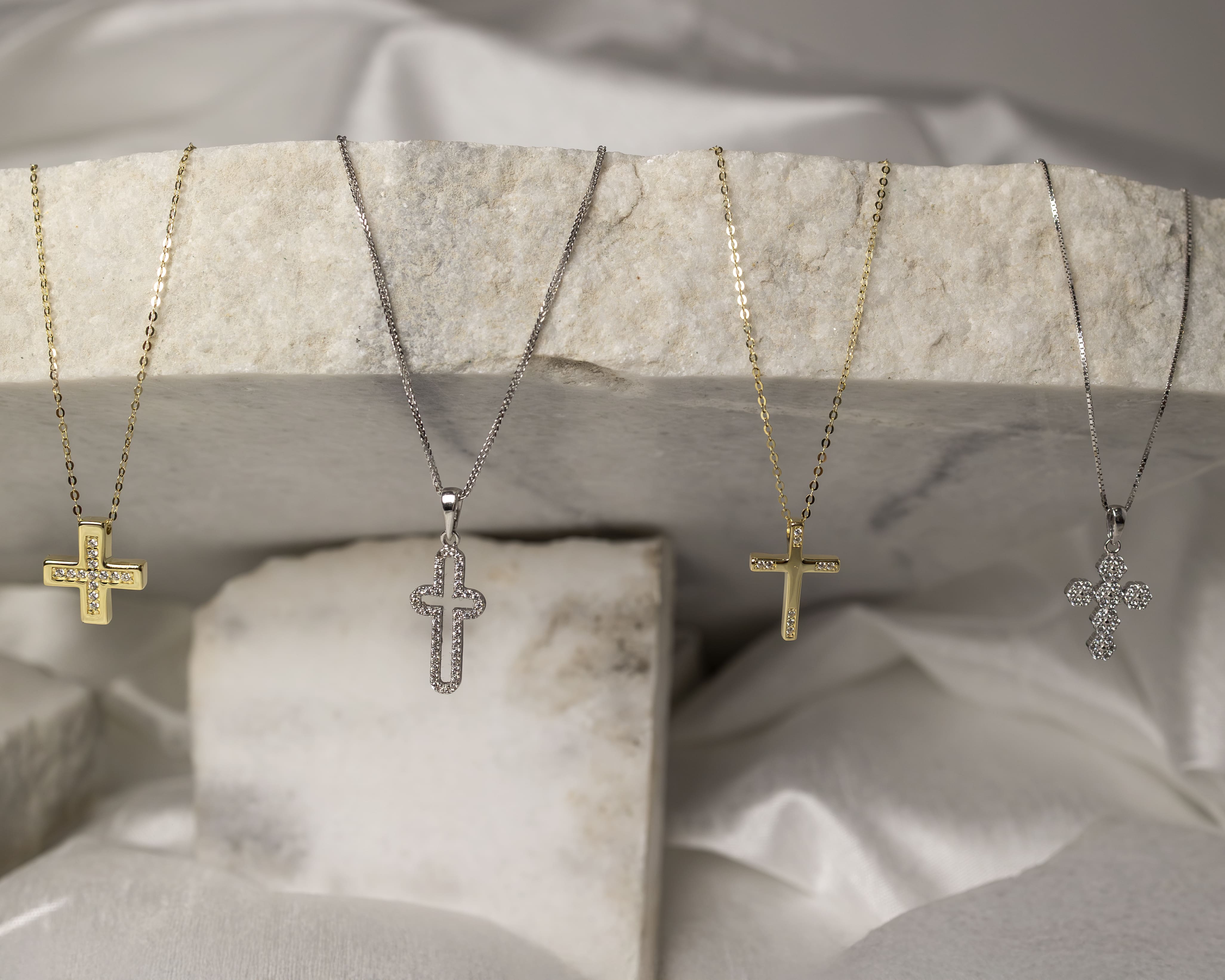 Four cross necklaces with gold and silver chains, each adorned with small crystals, displayed on a textured stone surface.