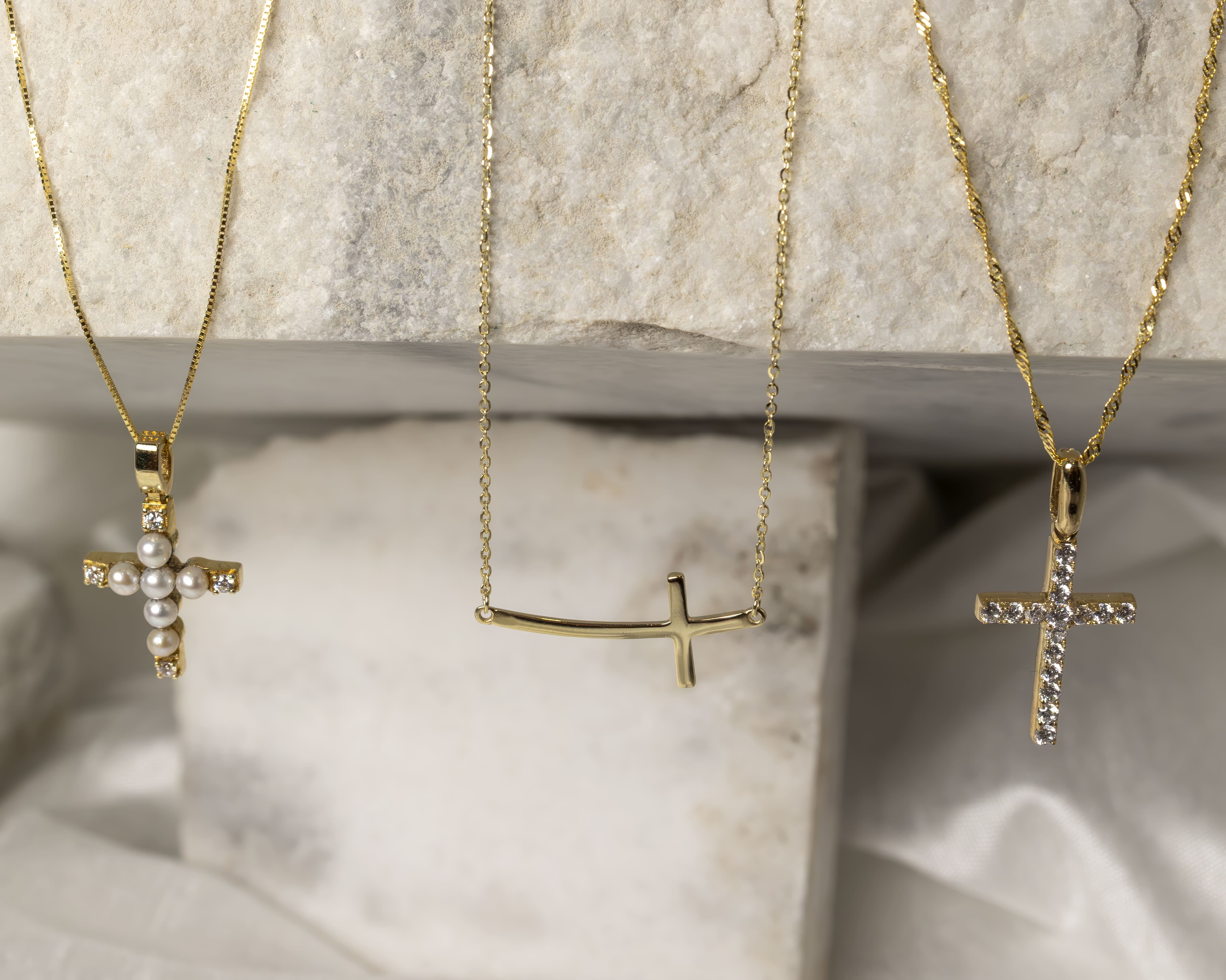 Three gold cross necklaces displayed on a light marble surface, featuring pearl, plain, and diamond designs.