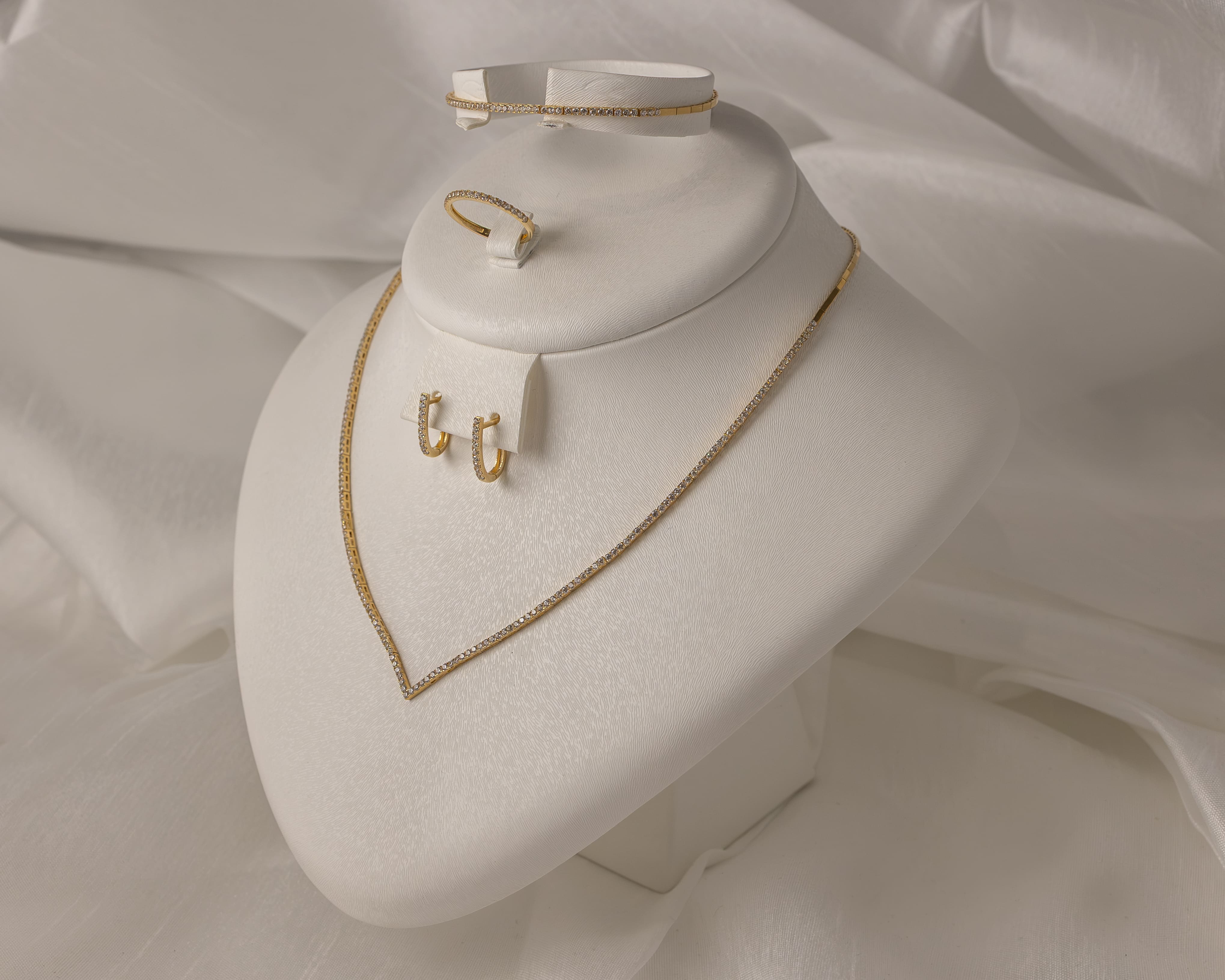 Gold jewelry set displayed on white stand, including a delicate V-shaped necklace, small hoop earrings, a ring, and a bracelet, all adorned with small diamonds.