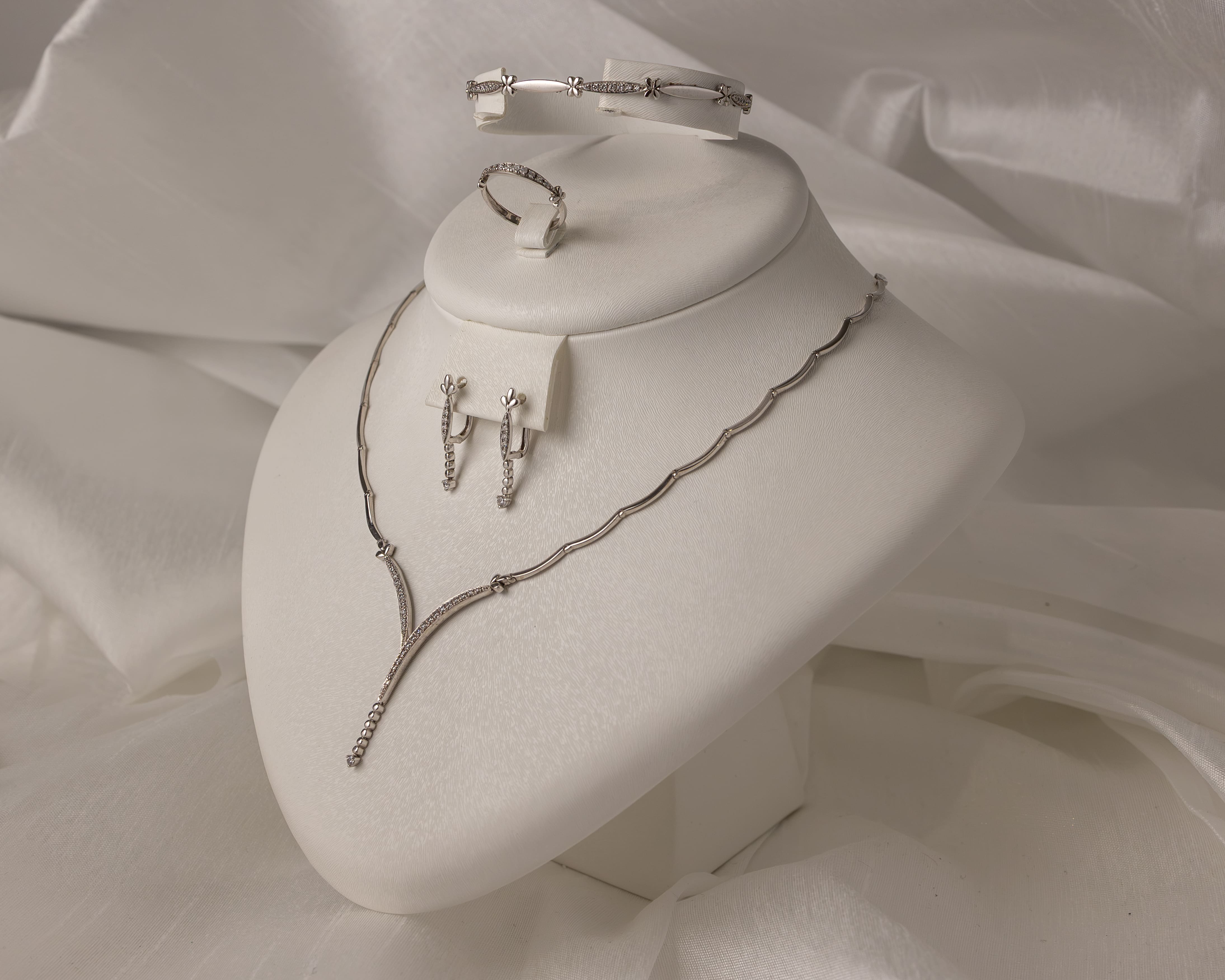 White jewelry display with a silver necklace, matching earrings, a ring, and a bracelet set on a white fabric background.