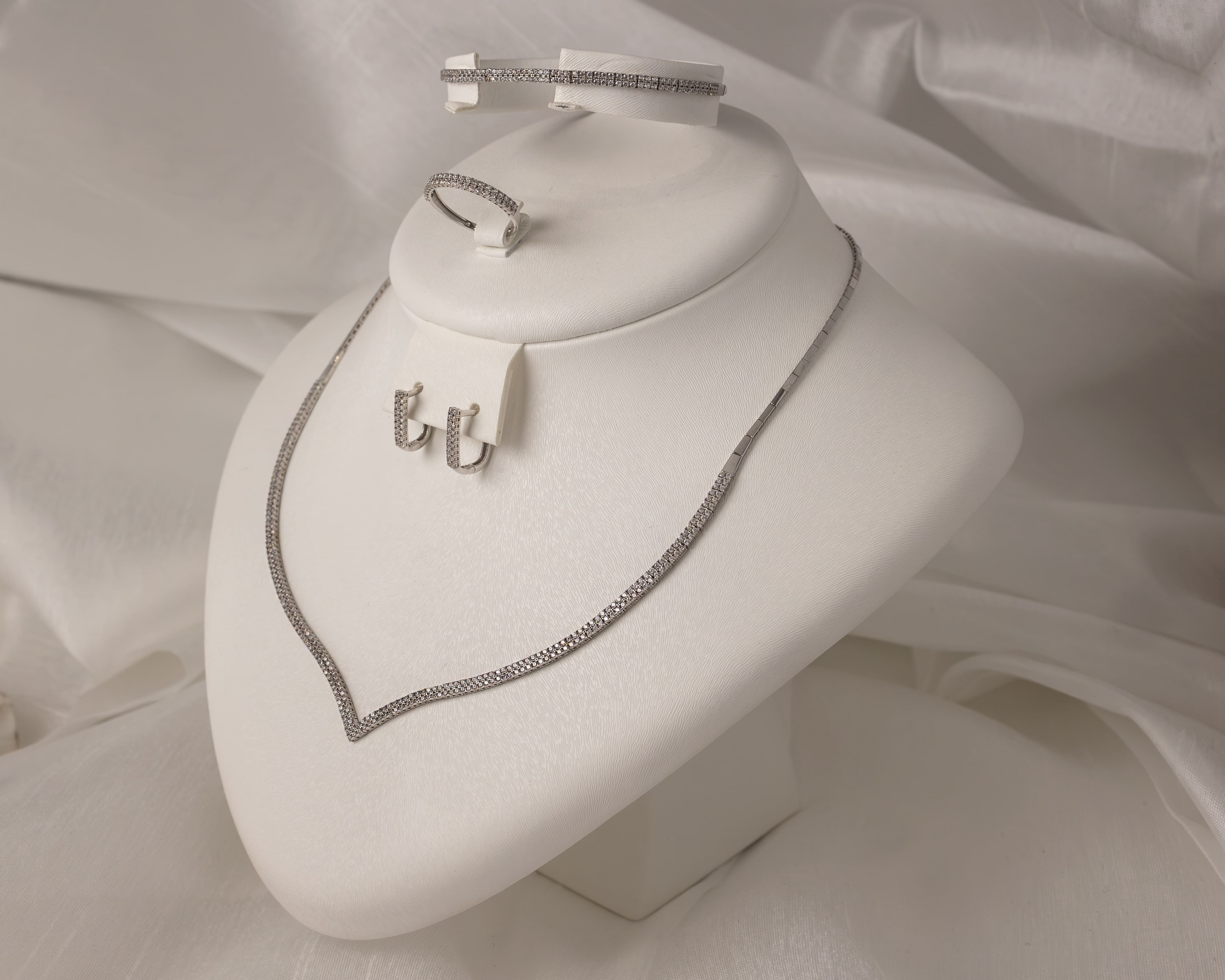 Set of silver jewelry with sparkling stones including a necklace, hoop earrings, a bracelet, and a ring displayed on a white stand.