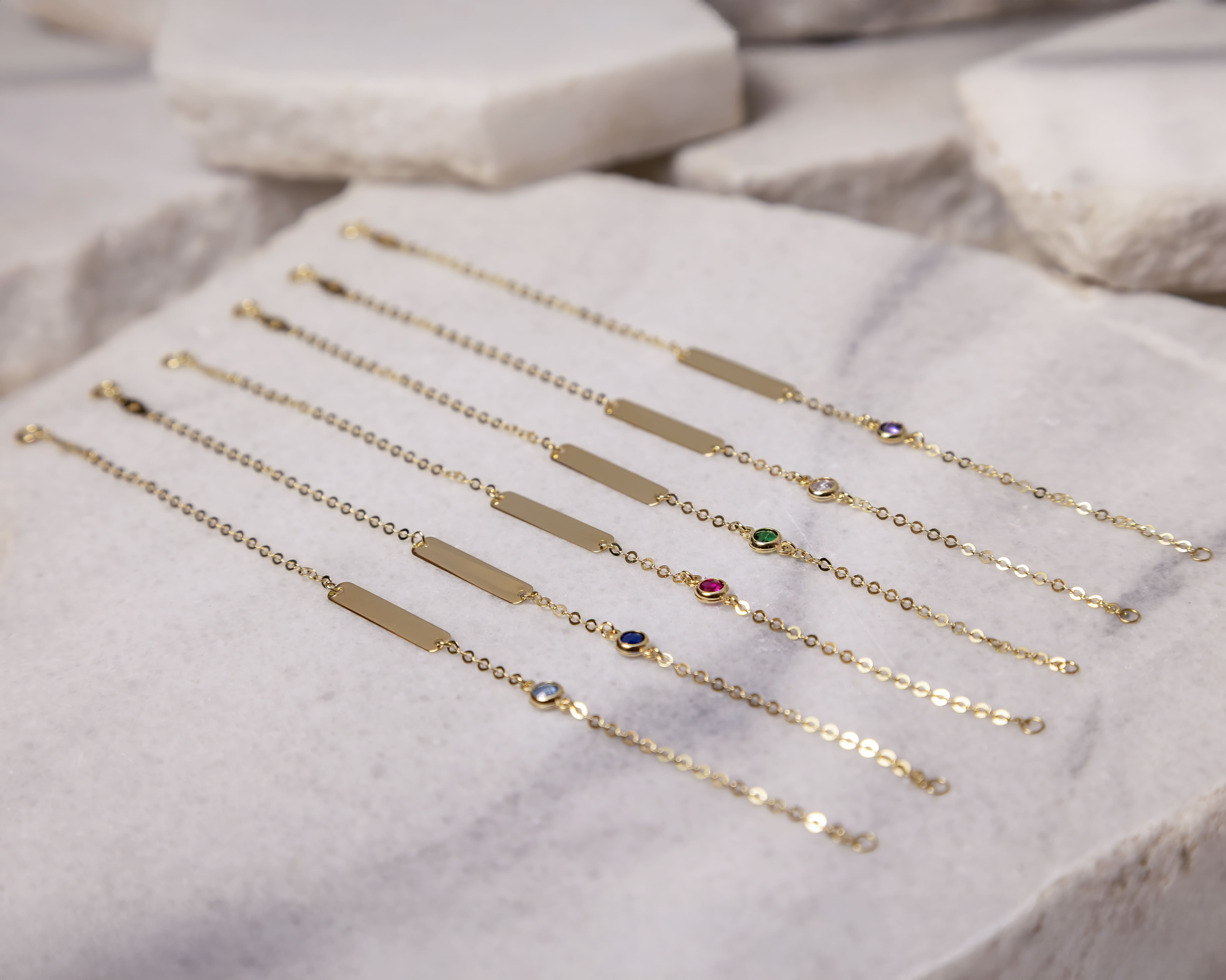 Six delicate gold chain bracelets with rectangular plates and small colorful gemstones, displayed on a stone surface.