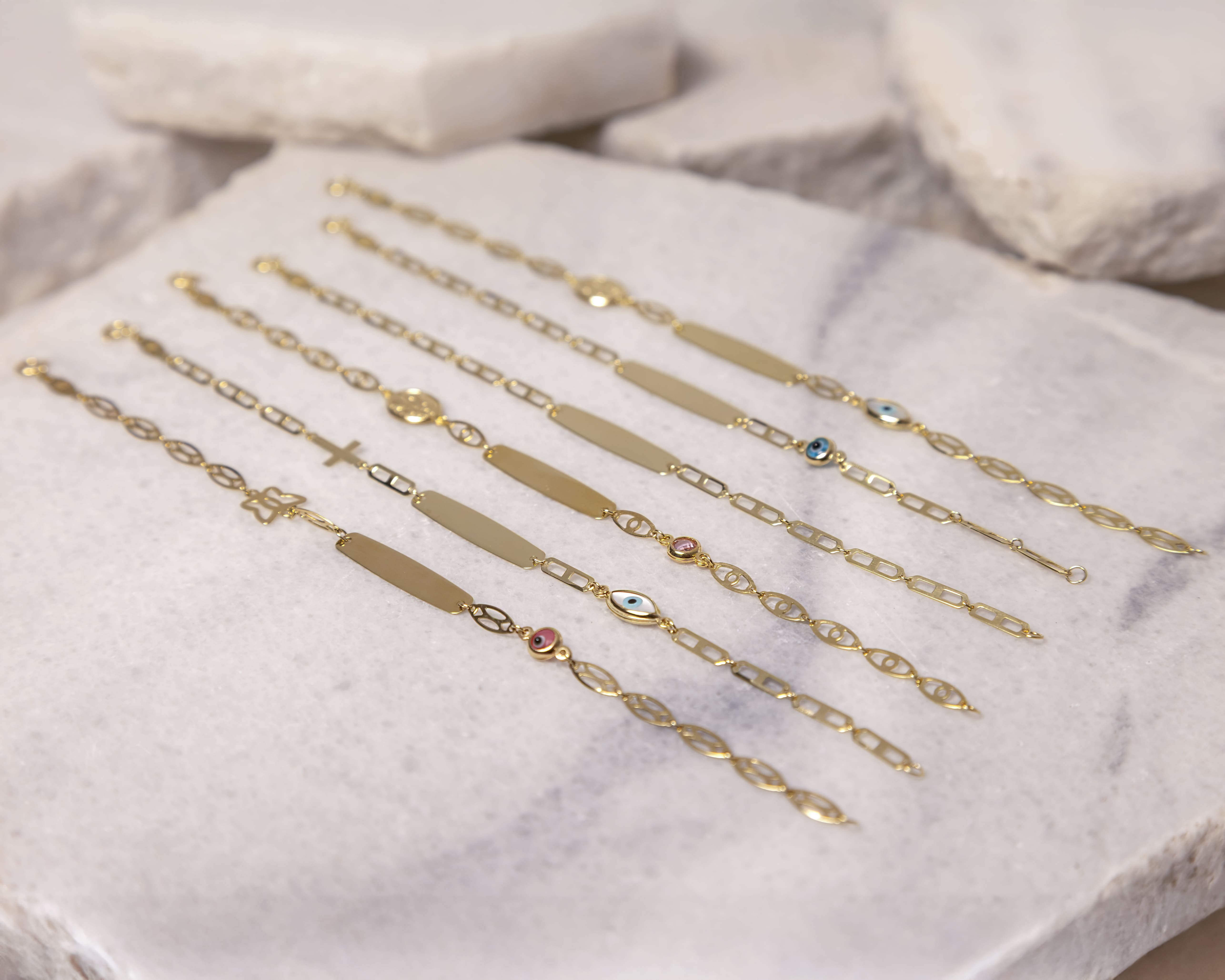 Six gold chain bracelets with various pendants and blank nameplates displayed on a white stone surface.