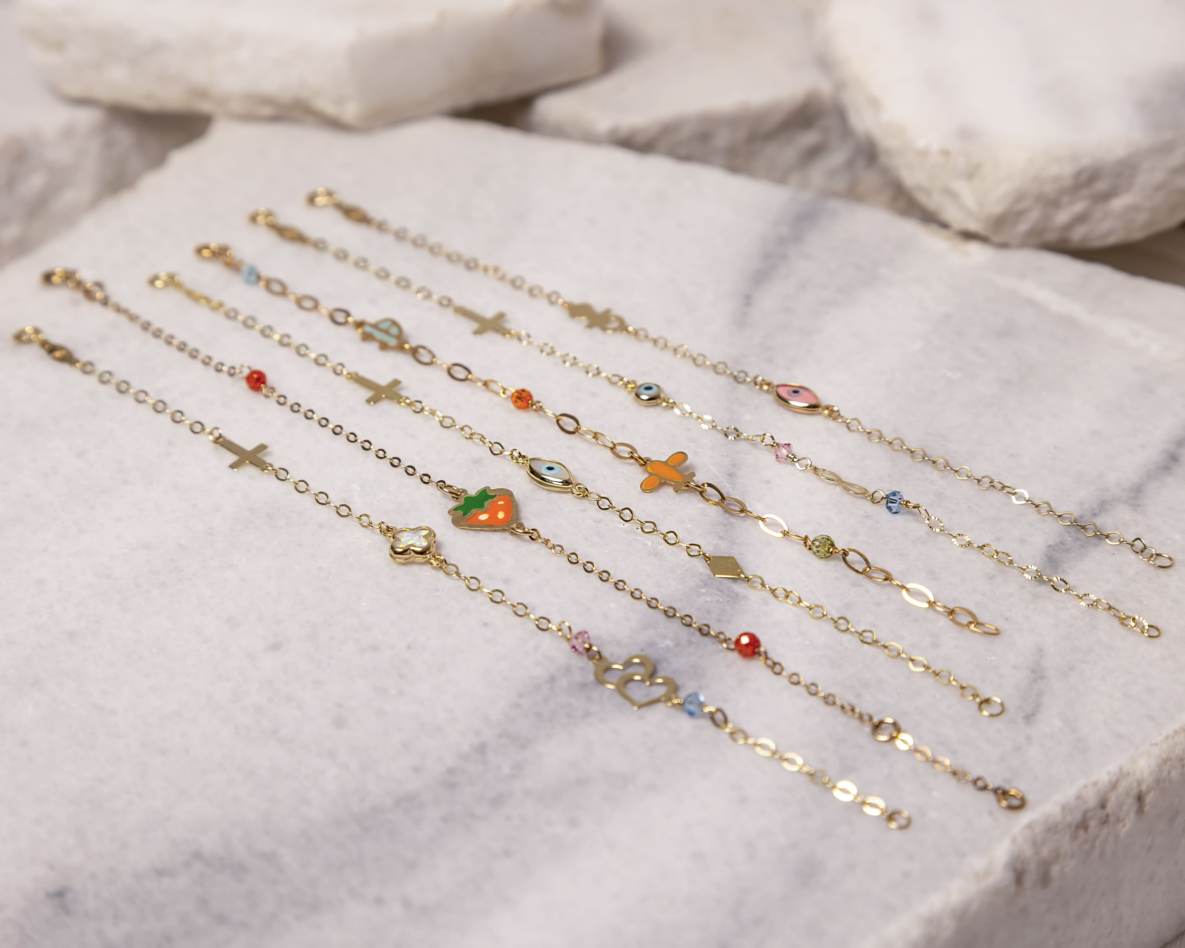 Seven delicate gold chain bracelets with various charms including crosses, a strawberry, an evil eye, a butterfly, and small colored beads laid out on a light stone surface.
