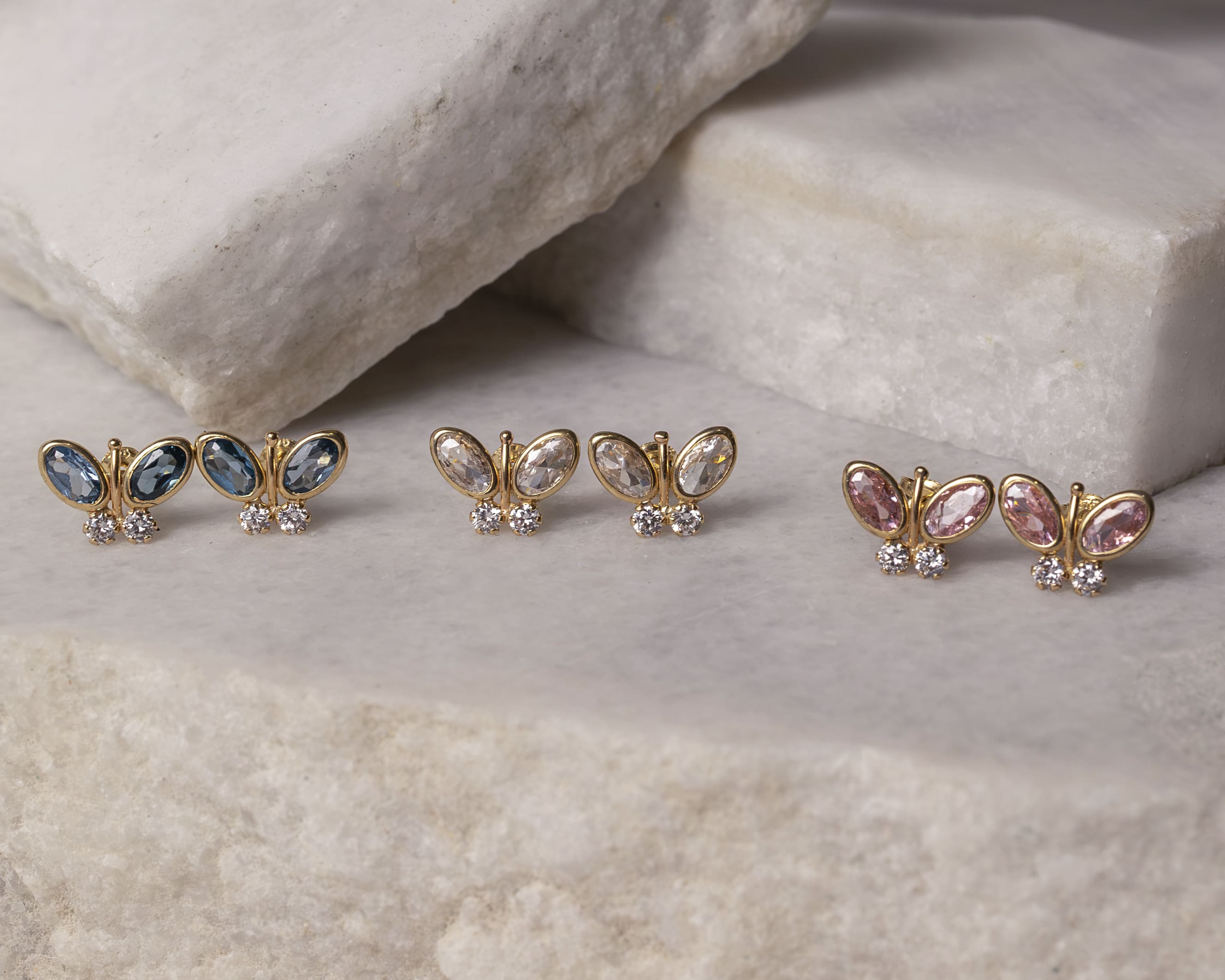 Three pairs of gold butterfly-shaped earrings with blue, clear, and pink gemstones on a white marble surface.