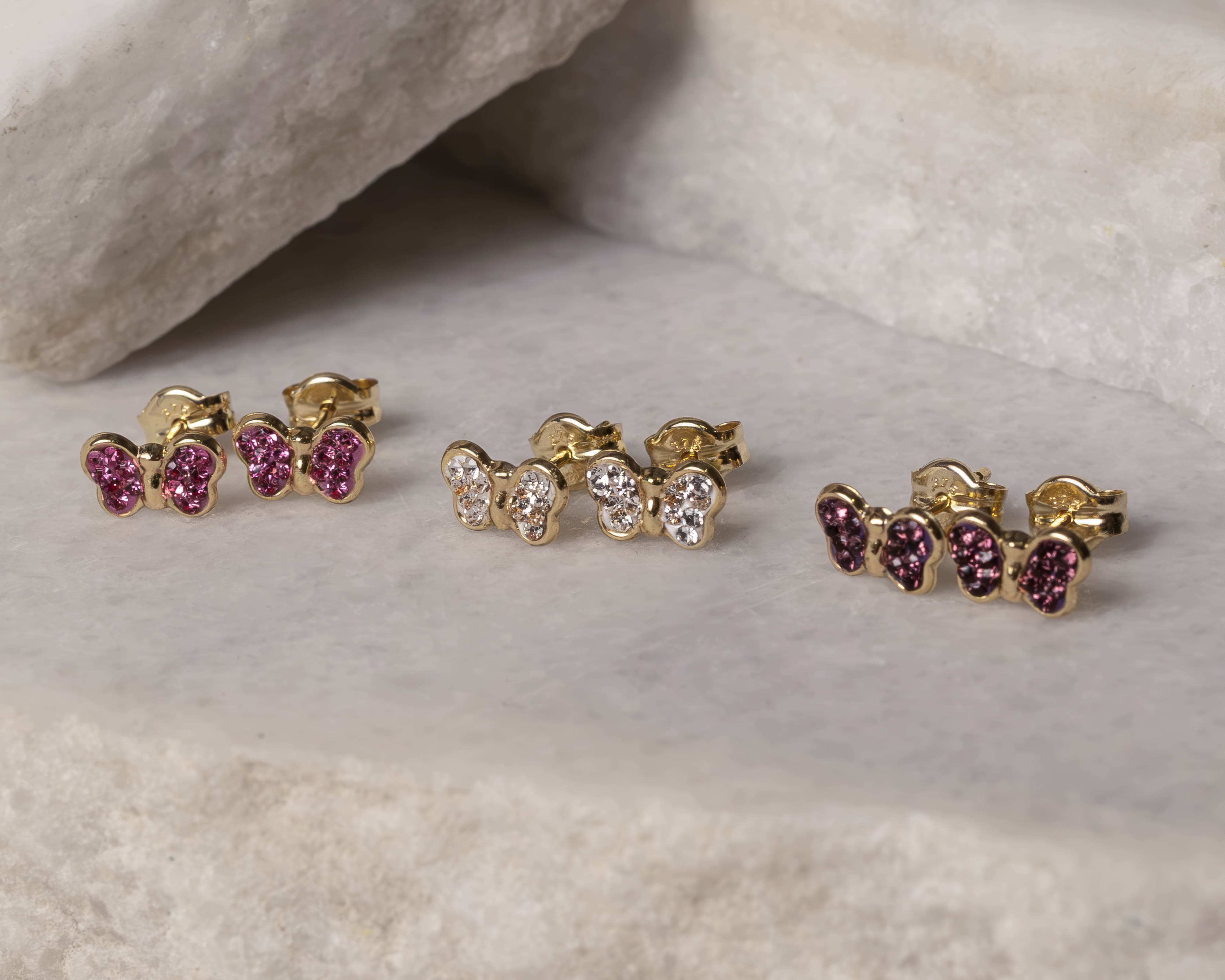 Three pairs of gold butterfly-shaped stud earrings with pink and clear gemstones on a marble surface.