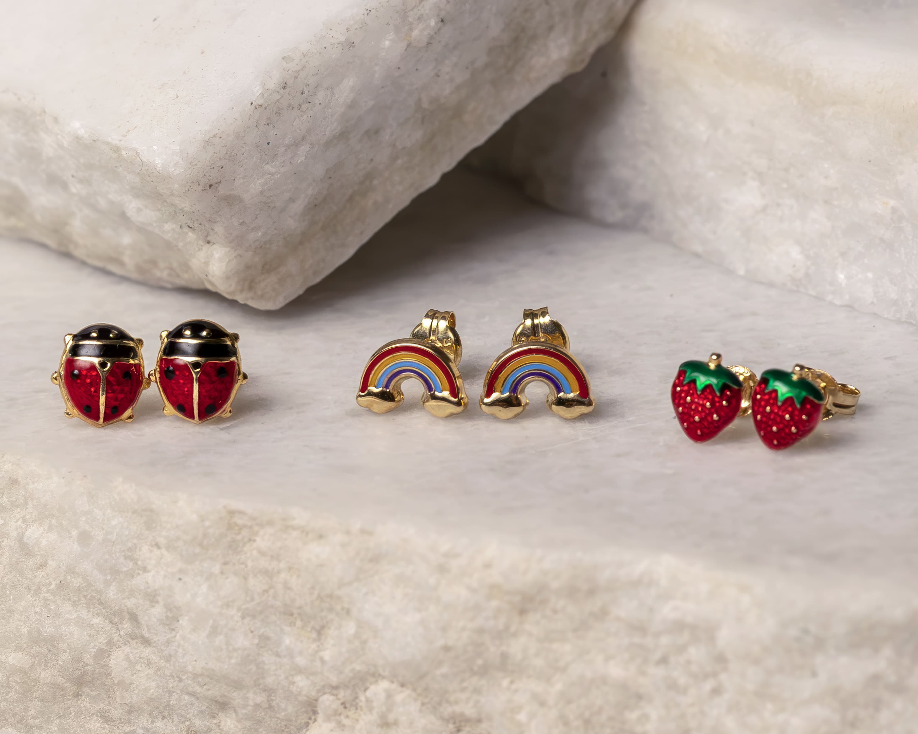 Three pairs of small gold-colored stud earrings shaped like ladybugs, rainbows, and strawberries on a marble surface.