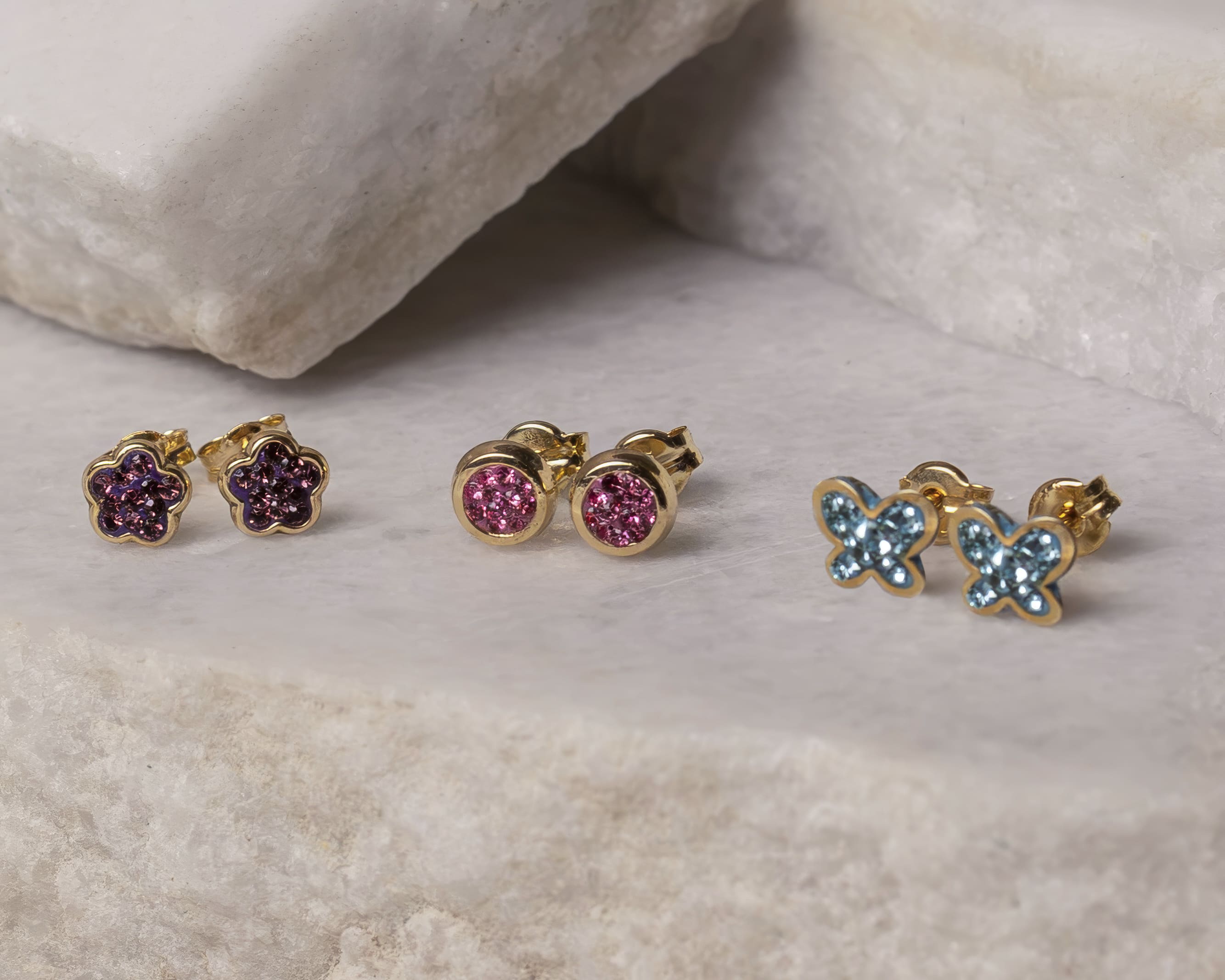 Three pairs of gold stud earrings with colored gemstones in flower, round, and butterfly shapes on a white stone surface.