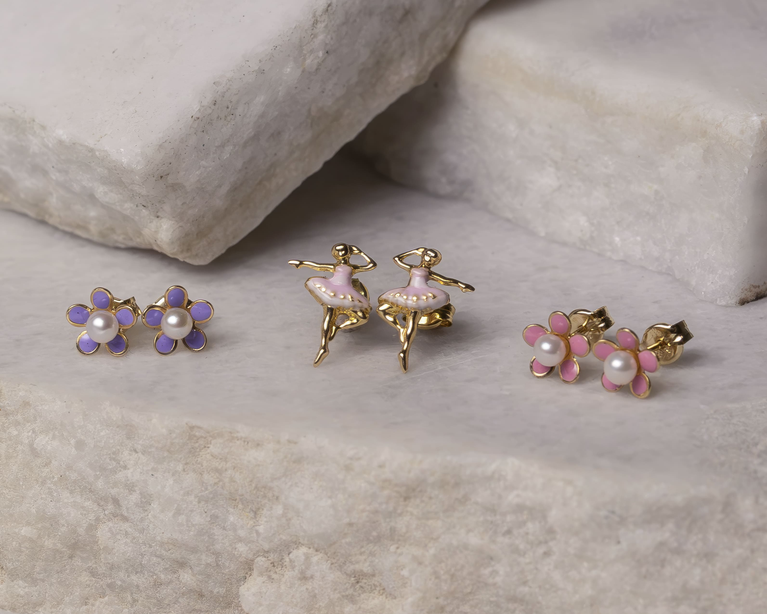 Three pairs of gold stud earrings on a stone surface: purple flower with pearl center, pink ballerina figurine, and pink flower with pearl center.