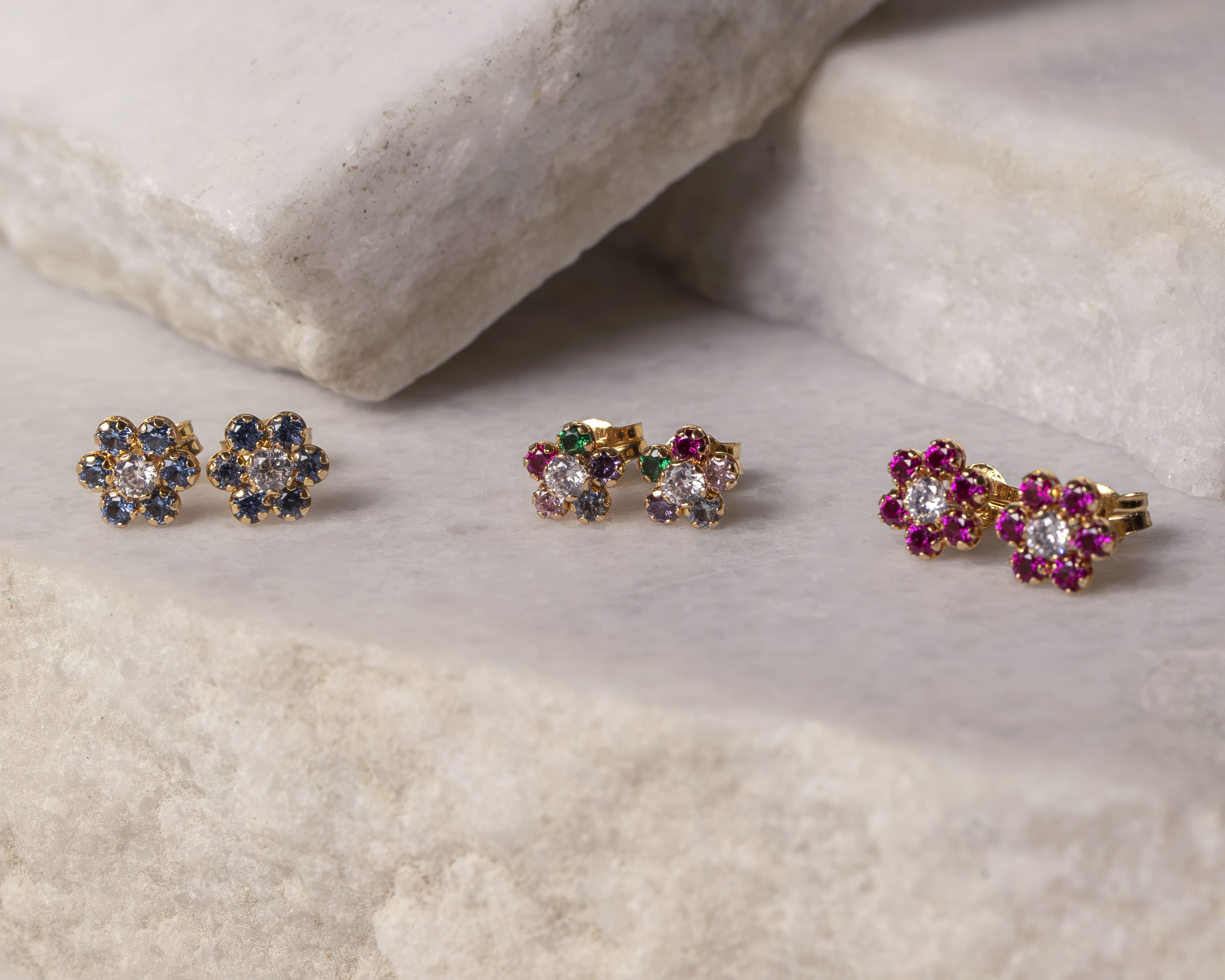 Three pairs of flower-shaped stud earrings with gold settings and colored gemstones on a marble surface.