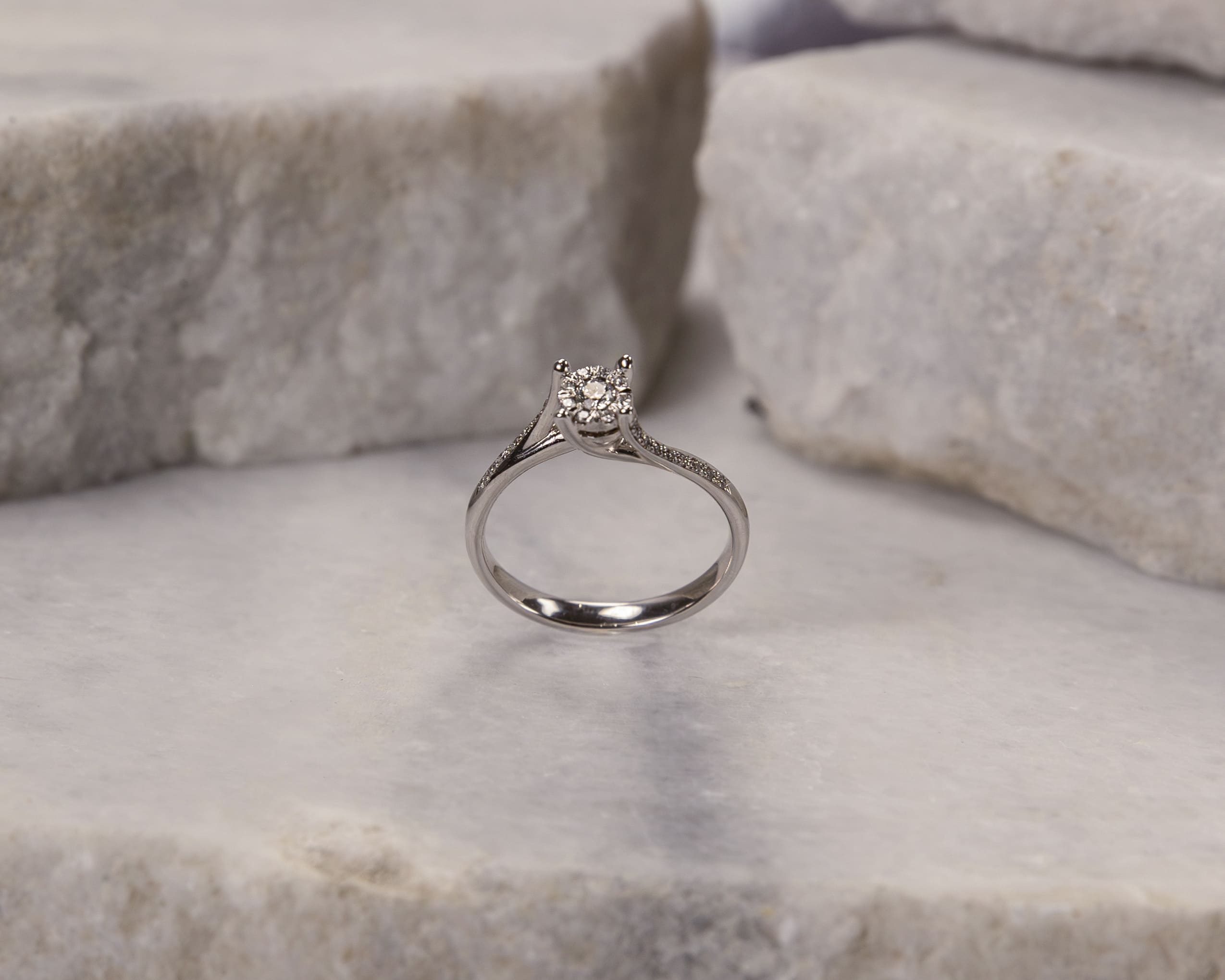 Silver ring with a cluster of small diamonds on a smooth marble surface with rough stone blocks in the background.