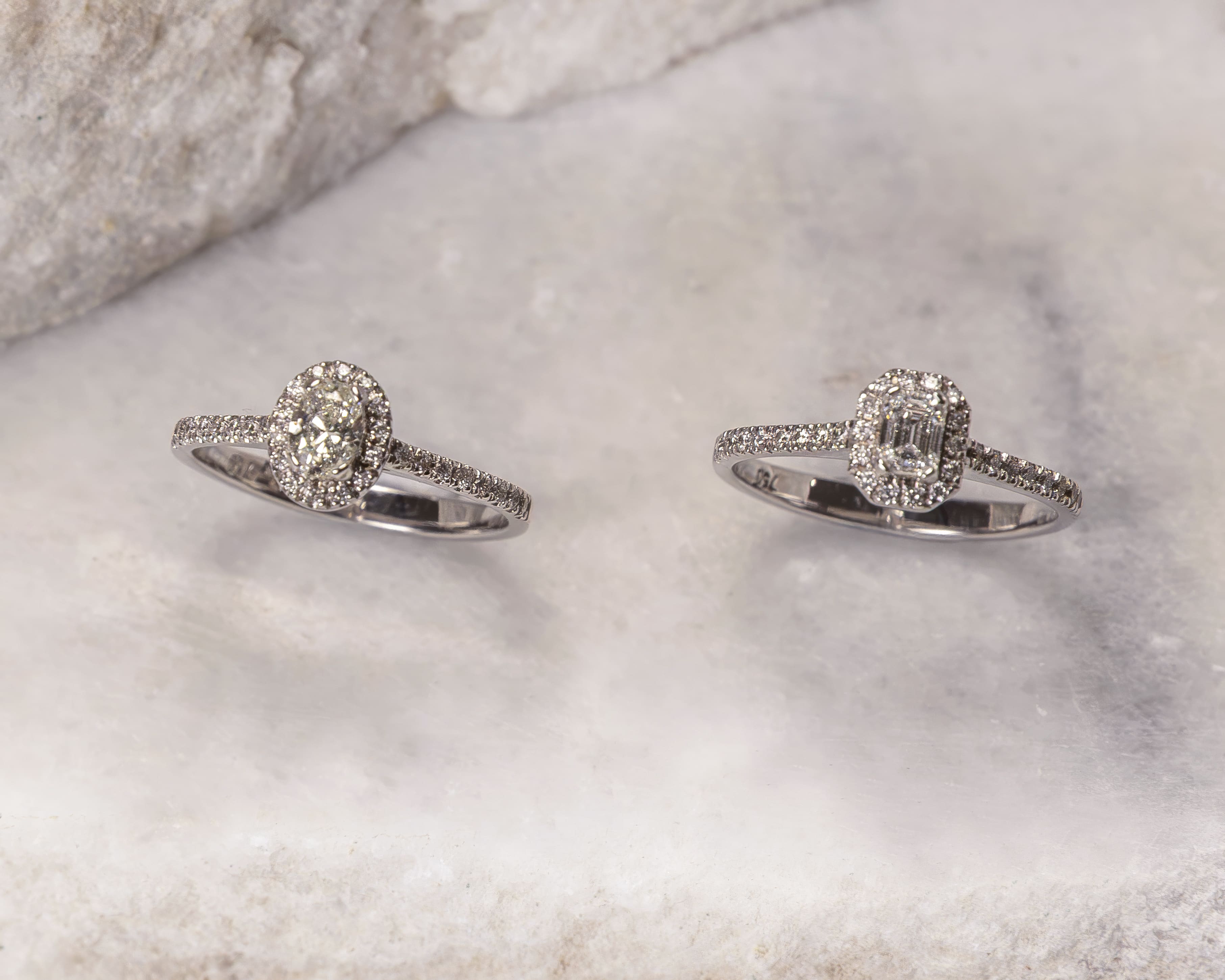 Two diamond rings on a marble surface, one with an oval center stone and the other with a rectangular center stone.