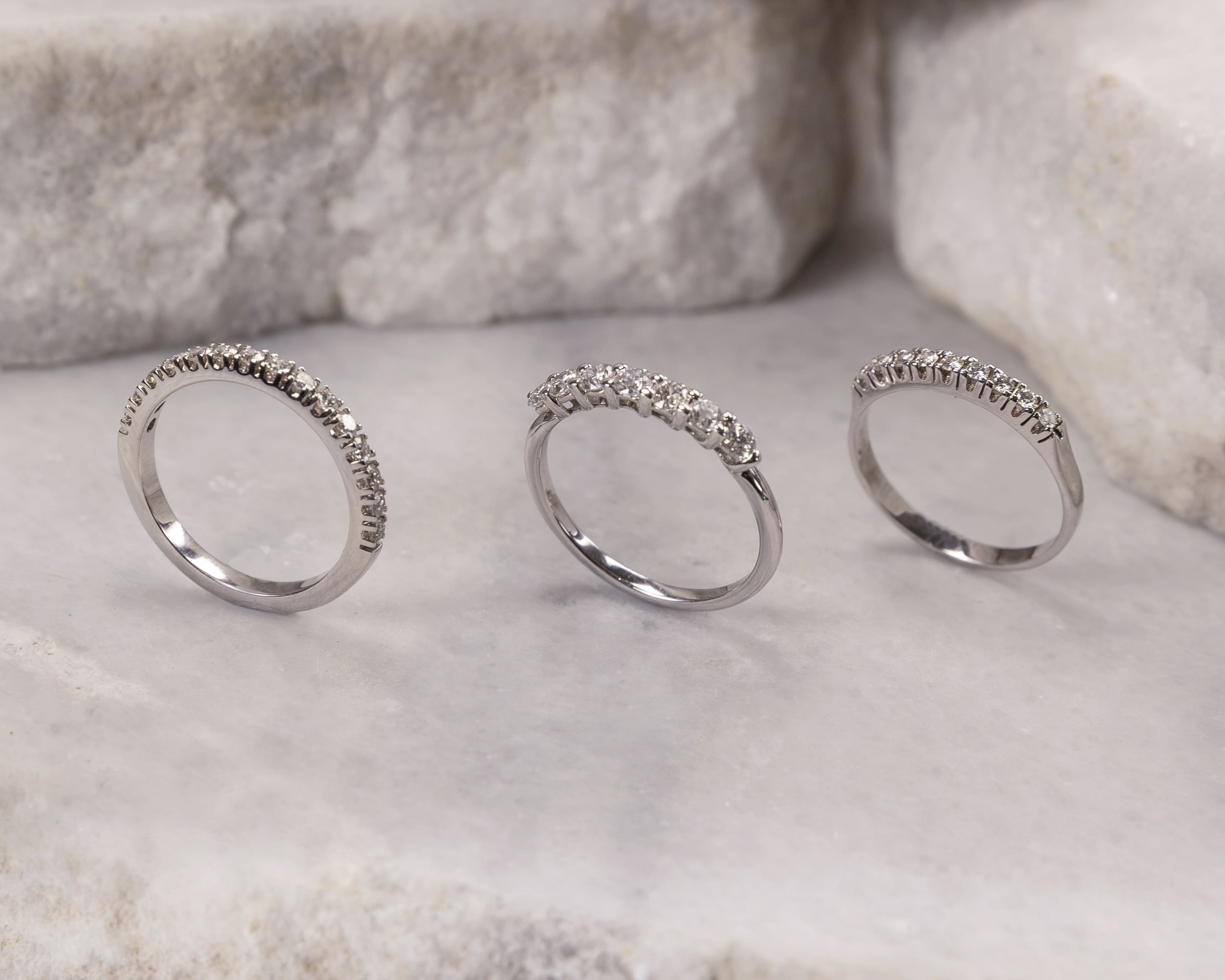 Three silver rings with small diamonds set in a row, placed on a light marble surface with large textured white stones in the background.