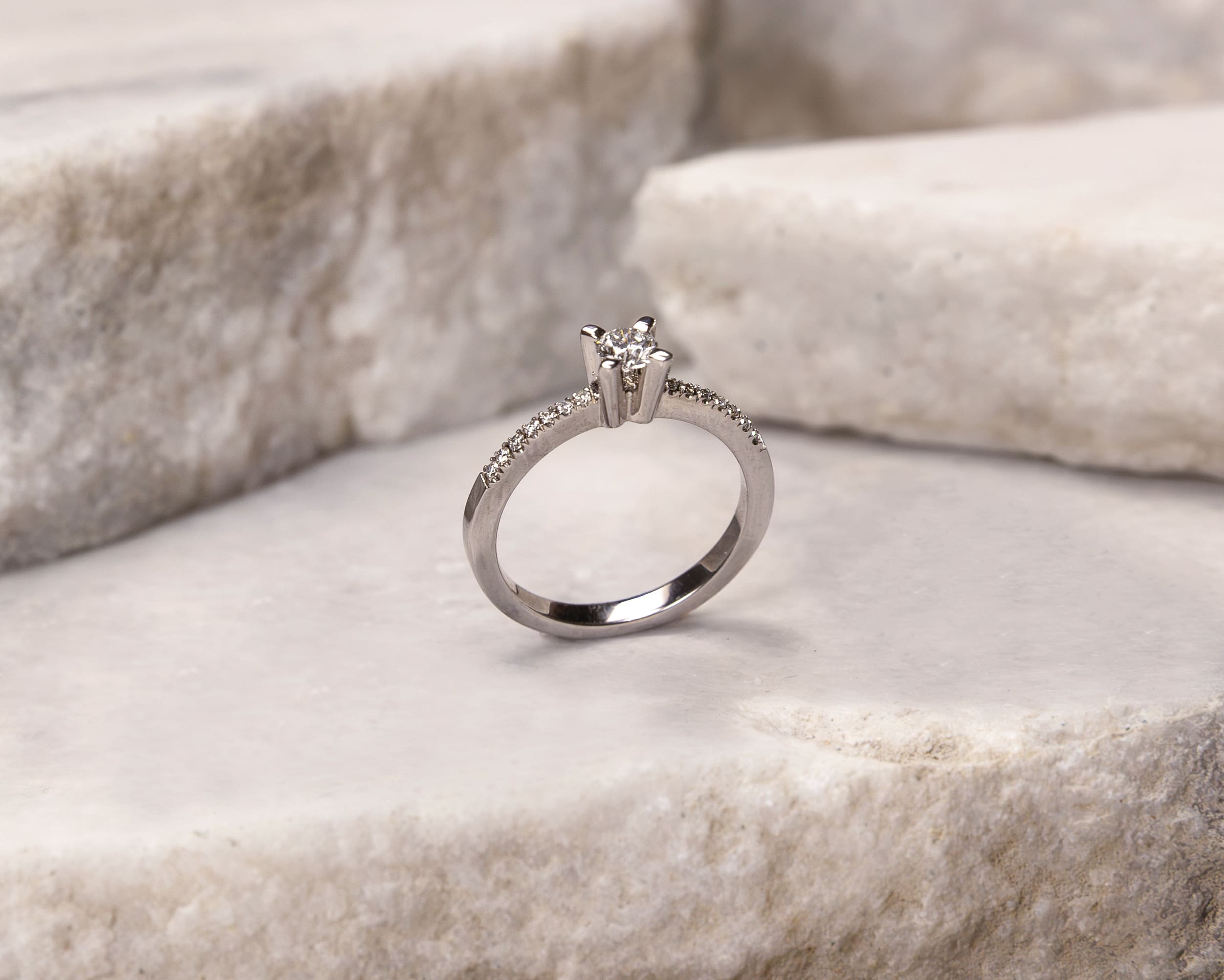 Silver ring with a central diamond and smaller diamonds along the band displayed on textured stone slabs.