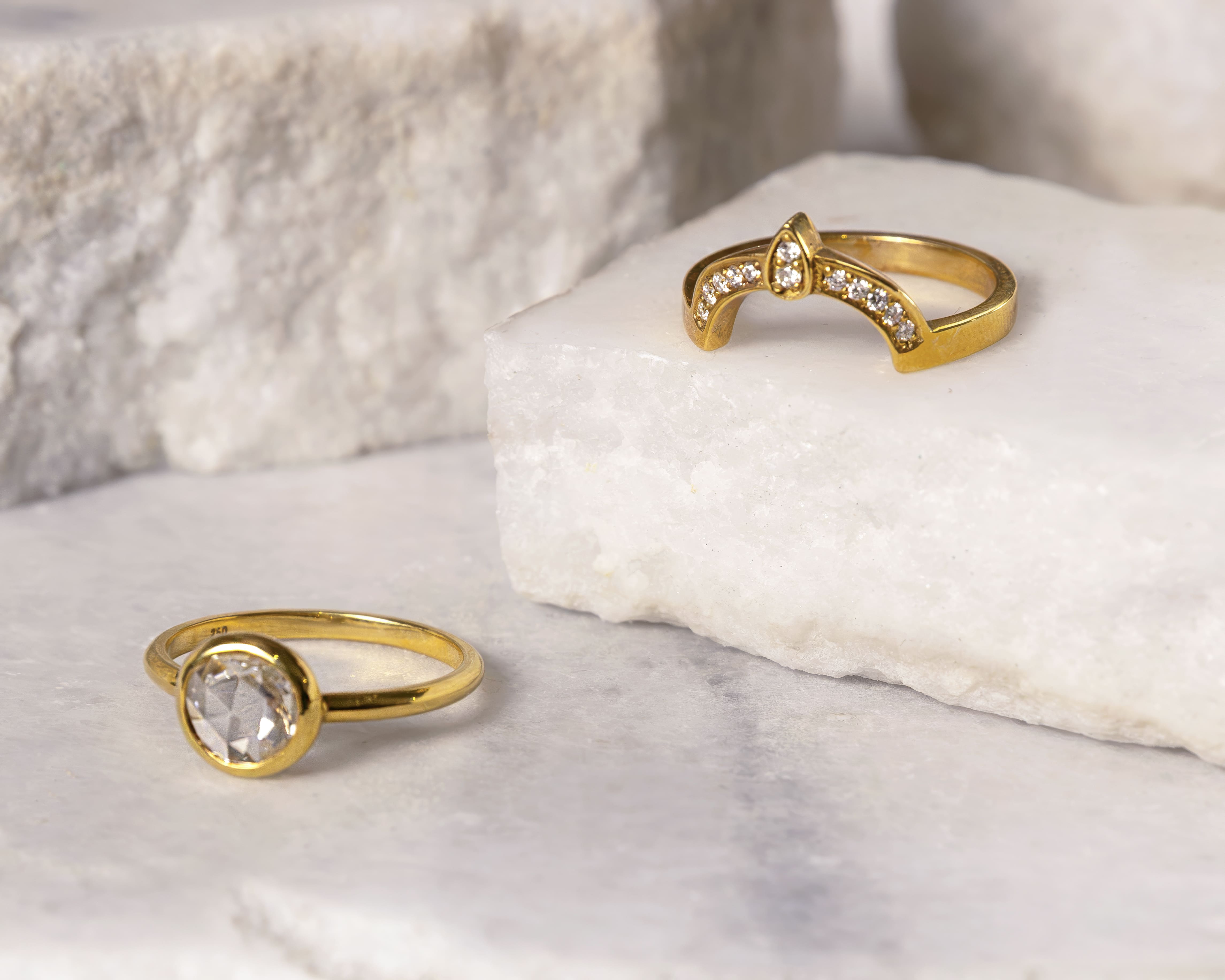 Two gold rings with clear gemstones placed on white marble and stone surfaces.