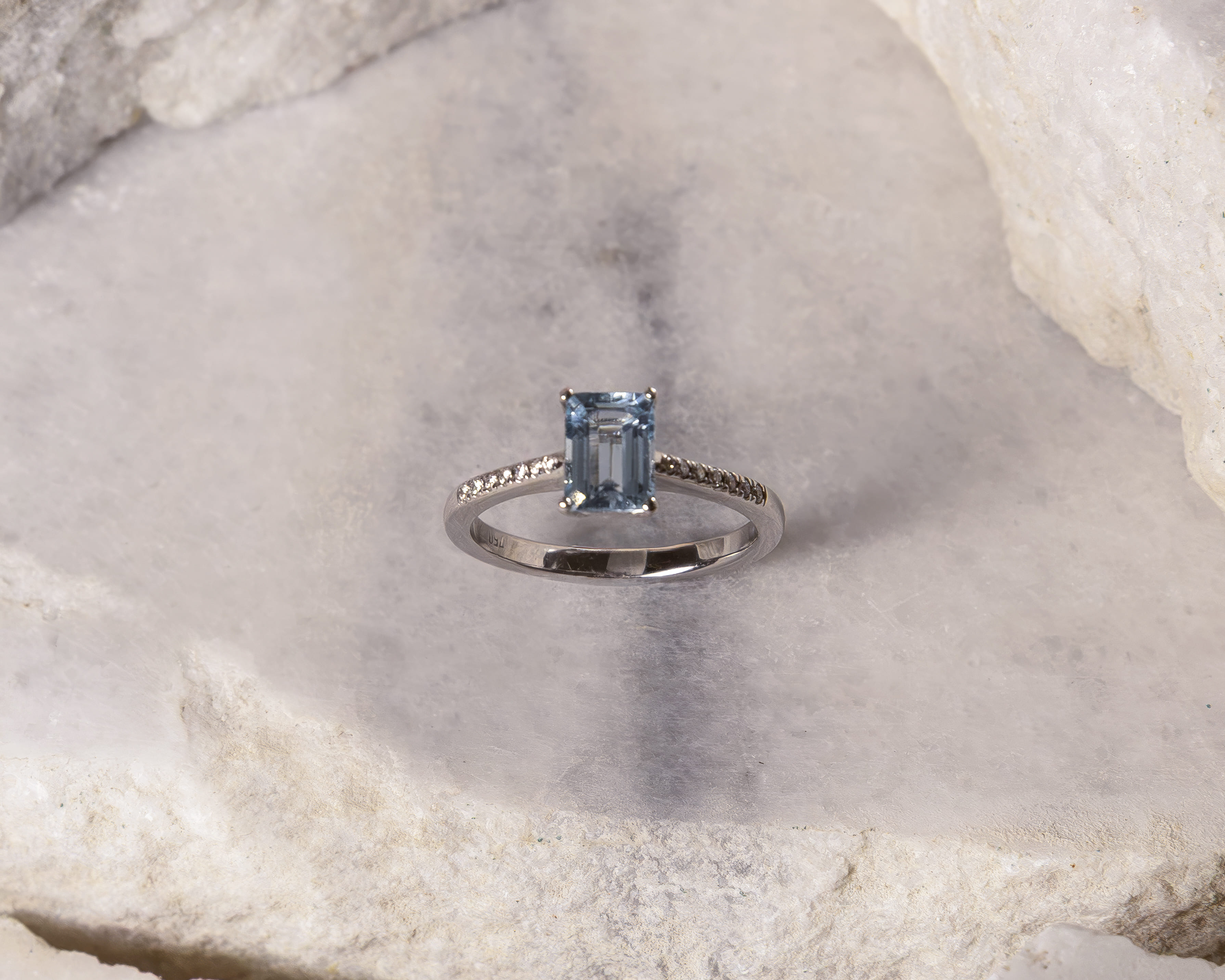 Silver ring with a rectangular light blue gemstone and small diamonds on the band, placed on a textured light stone surface.