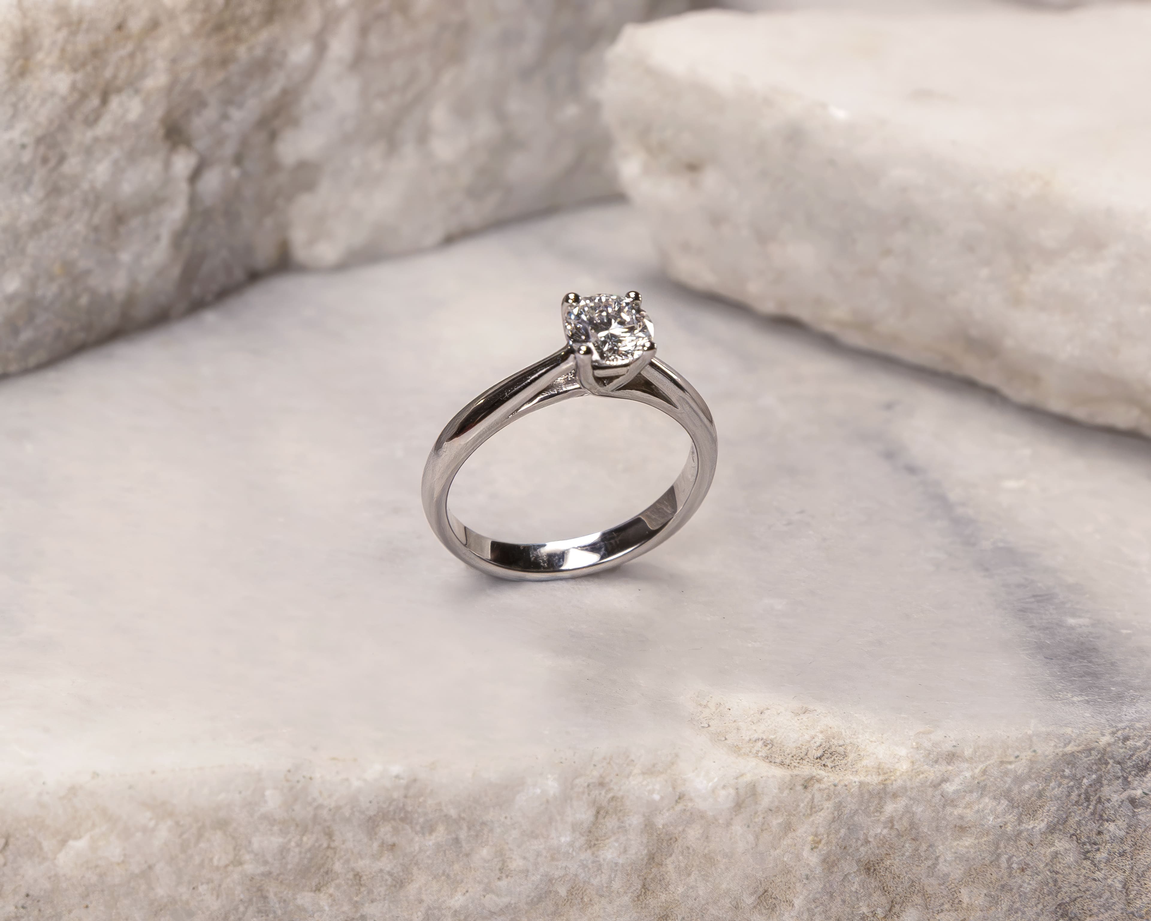Silver diamond solitaire ring displayed on textured light stone surface.