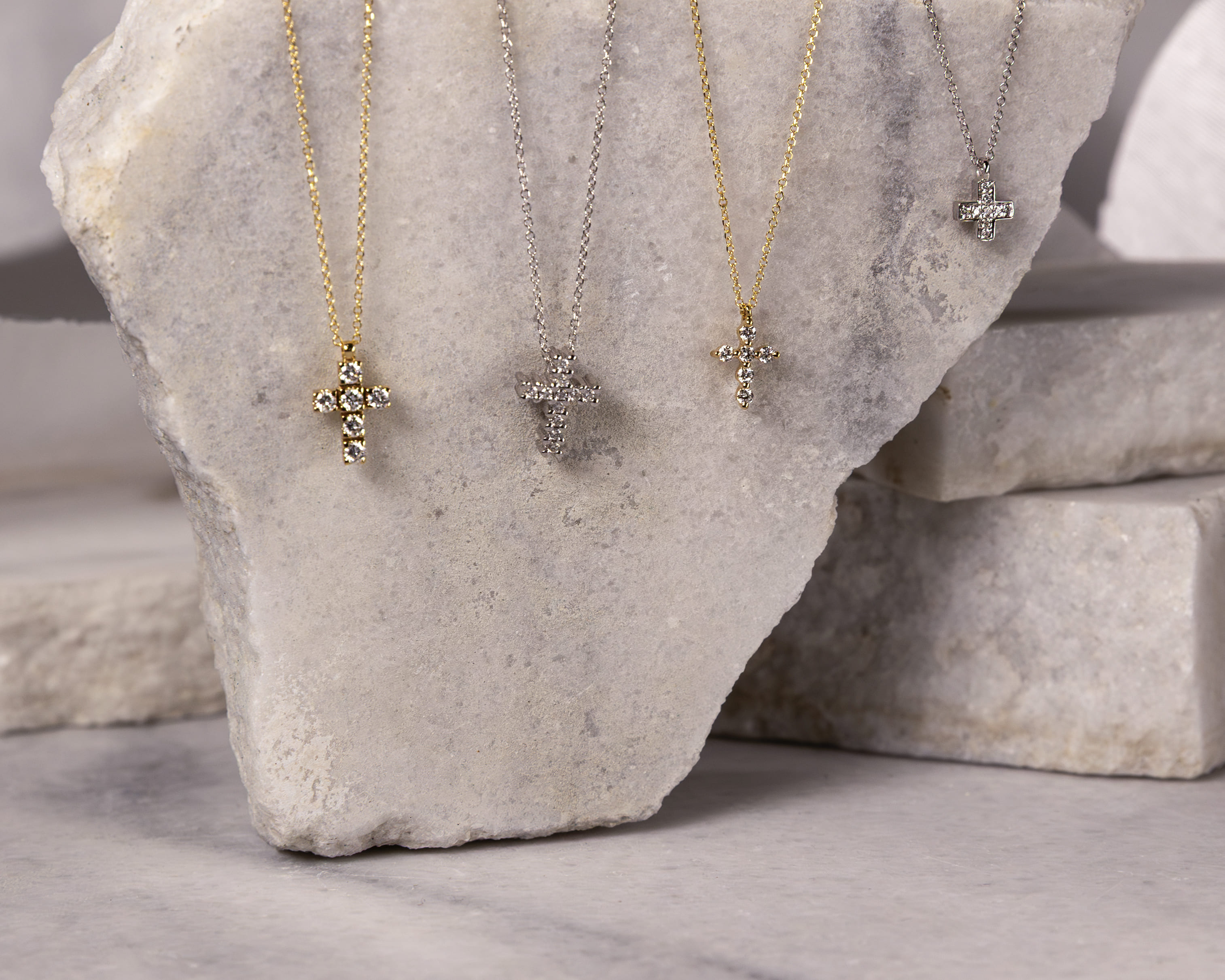Four cross-shaped diamond pendants on gold and silver chains displayed on a rough stone slab.