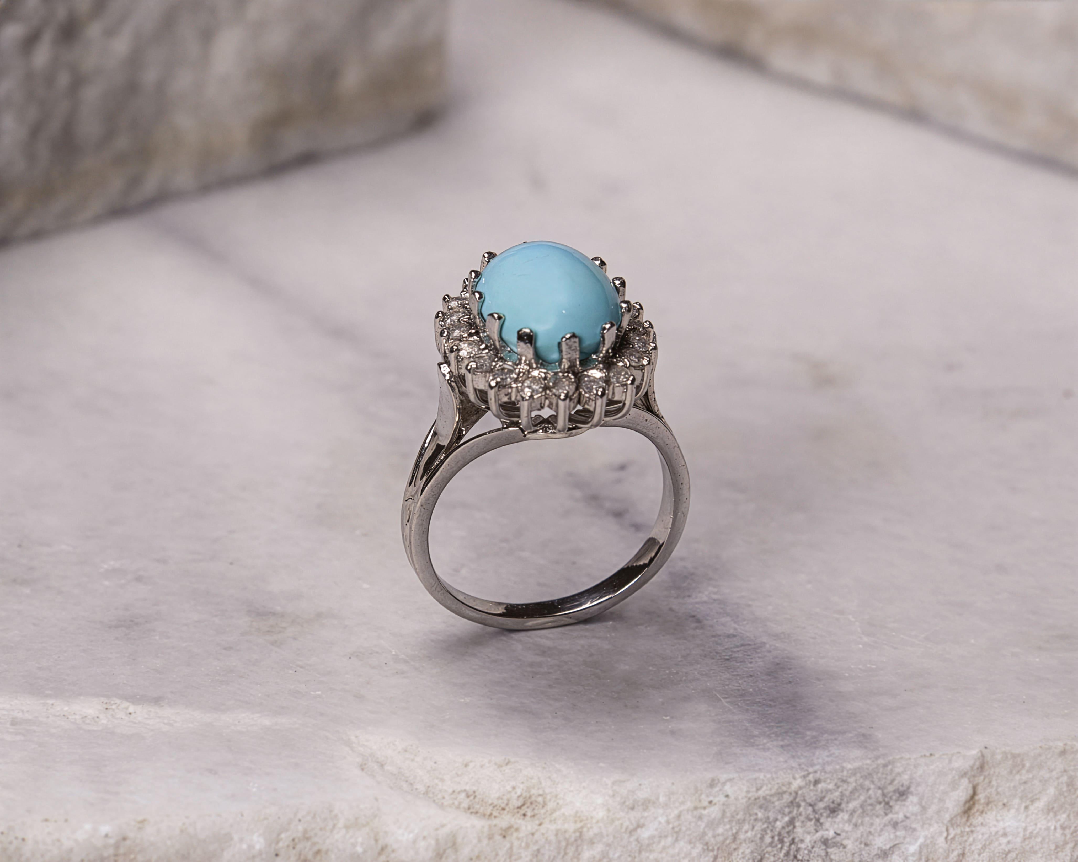 Silver ring with an oval light blue gemstone surrounded by small clear stones on a marble surface.