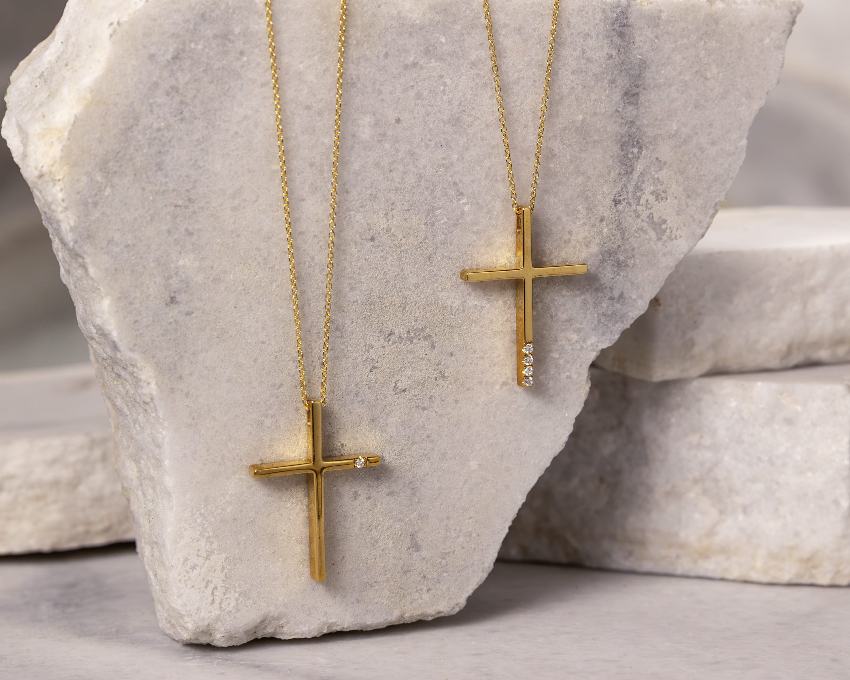 Two gold cross necklaces with diamonds on thin gold chains displayed on a textured gray stone surface.