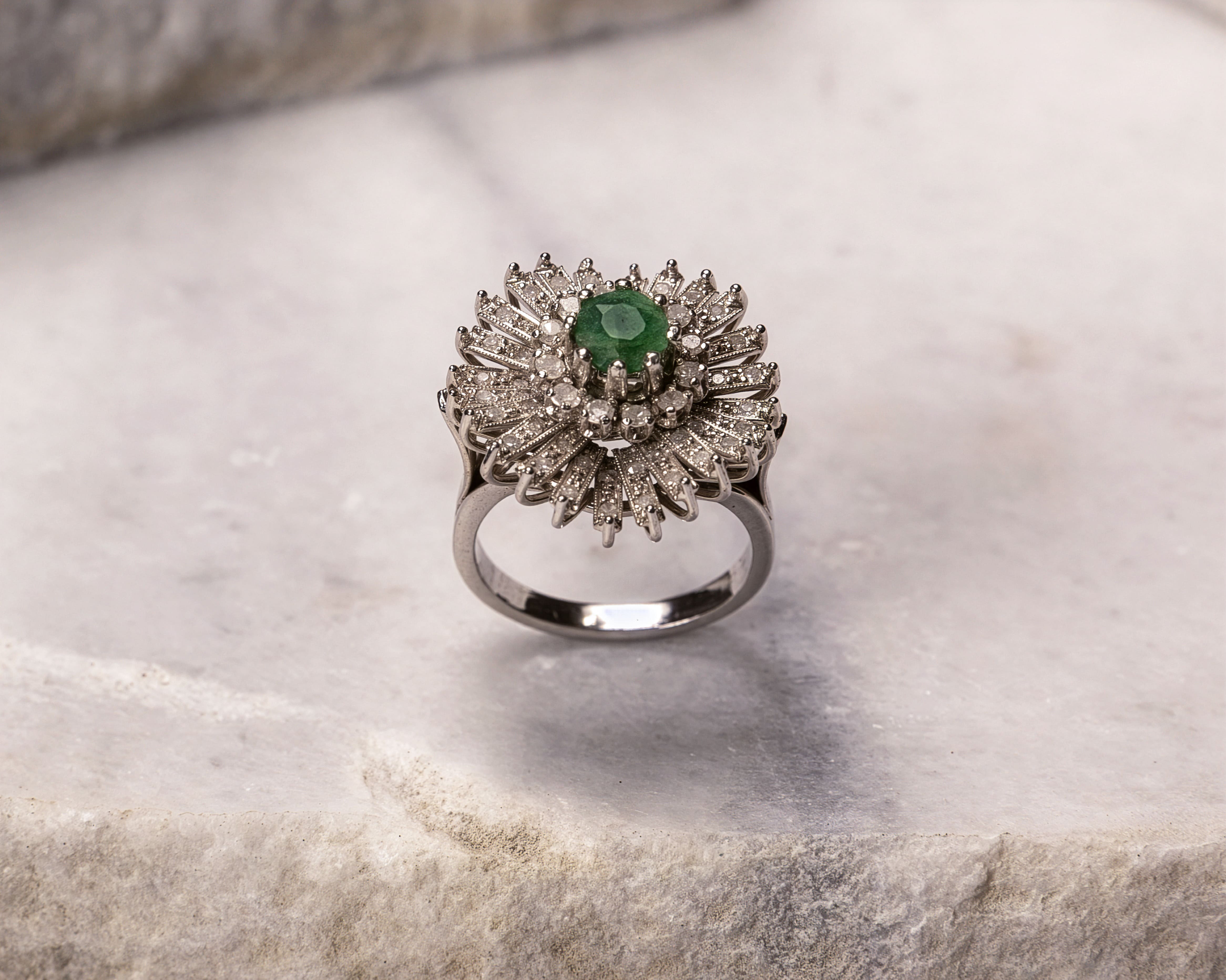 Silver ring with a central green gemstone surrounded by two layers of white diamonds arranged in a radiating pattern.