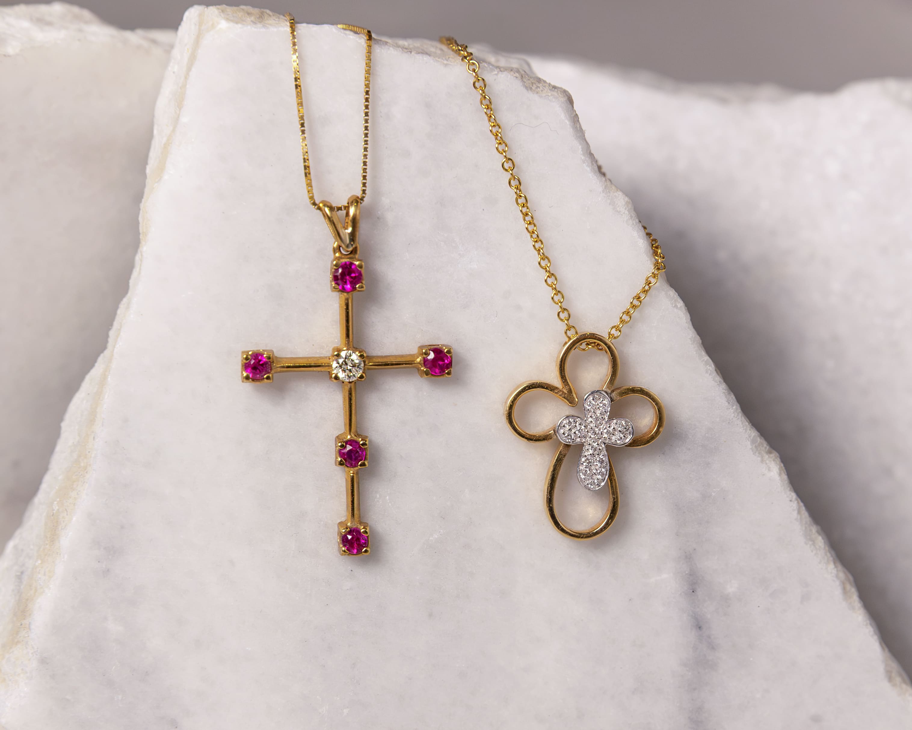 Two gold cross necklaces on a white marble surface, one with pink and white gemstones and the other with a looped design and small white gems.