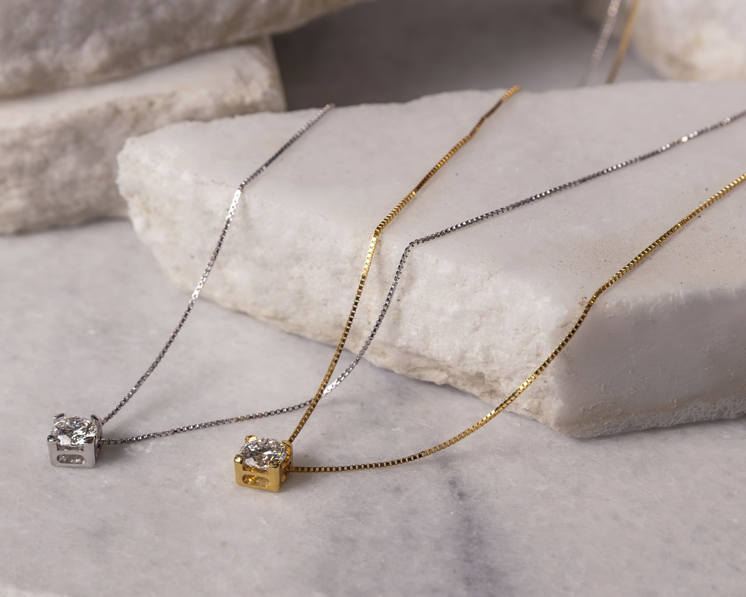 Two delicate necklaces with square pendants, one in gold and one in silver, each featuring a clear gemstone, displayed on white stone slabs.