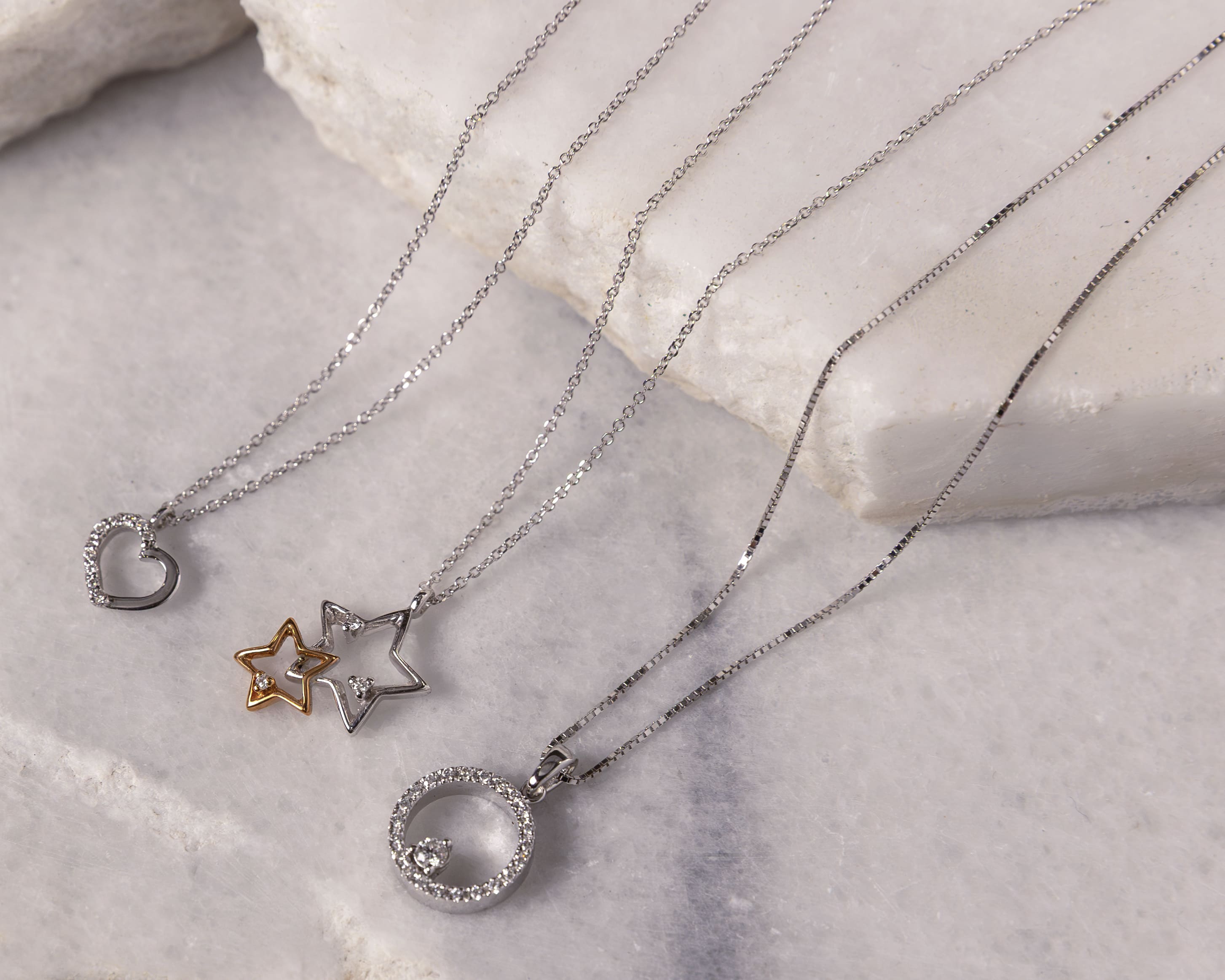 Three delicate silver necklaces with pendants shaped like a heart, double stars, and a circle with small gemstones on a marble surface.