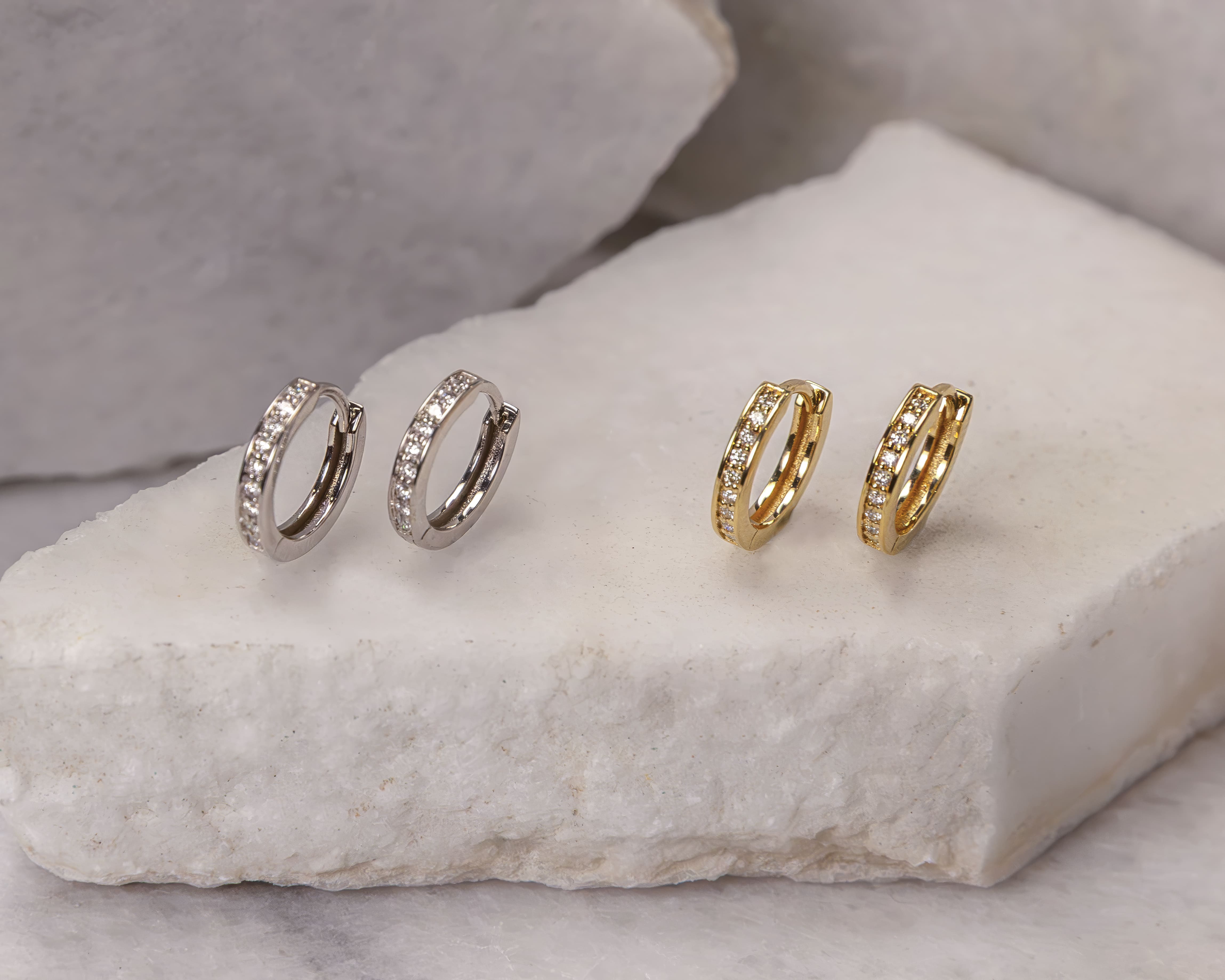 Two pairs of small hoop earrings with embedded stones, one pair in silver tone and one pair in gold tone, displayed on a white stone surface.