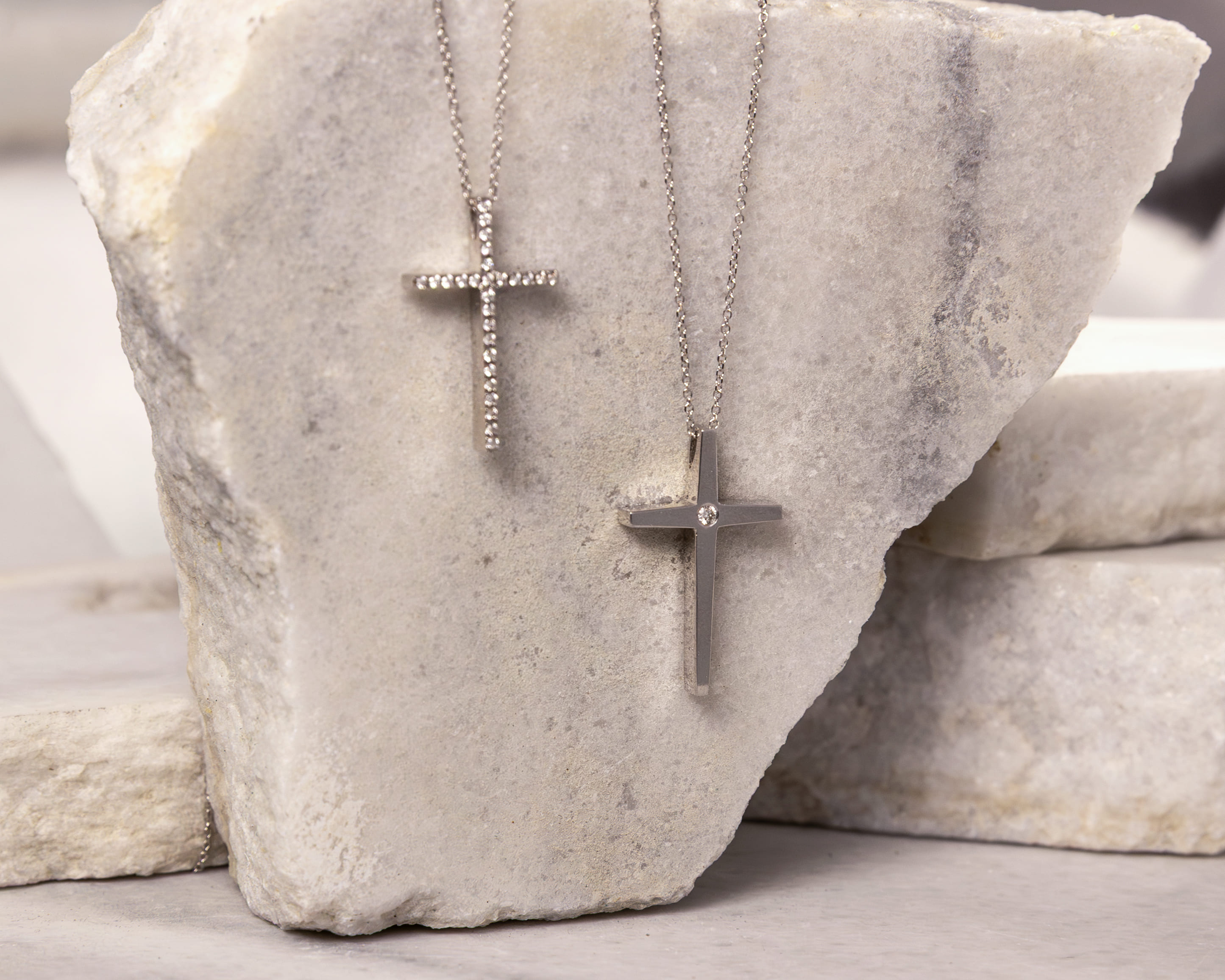 Two silver cross necklaces hanging on a textured white stone background.