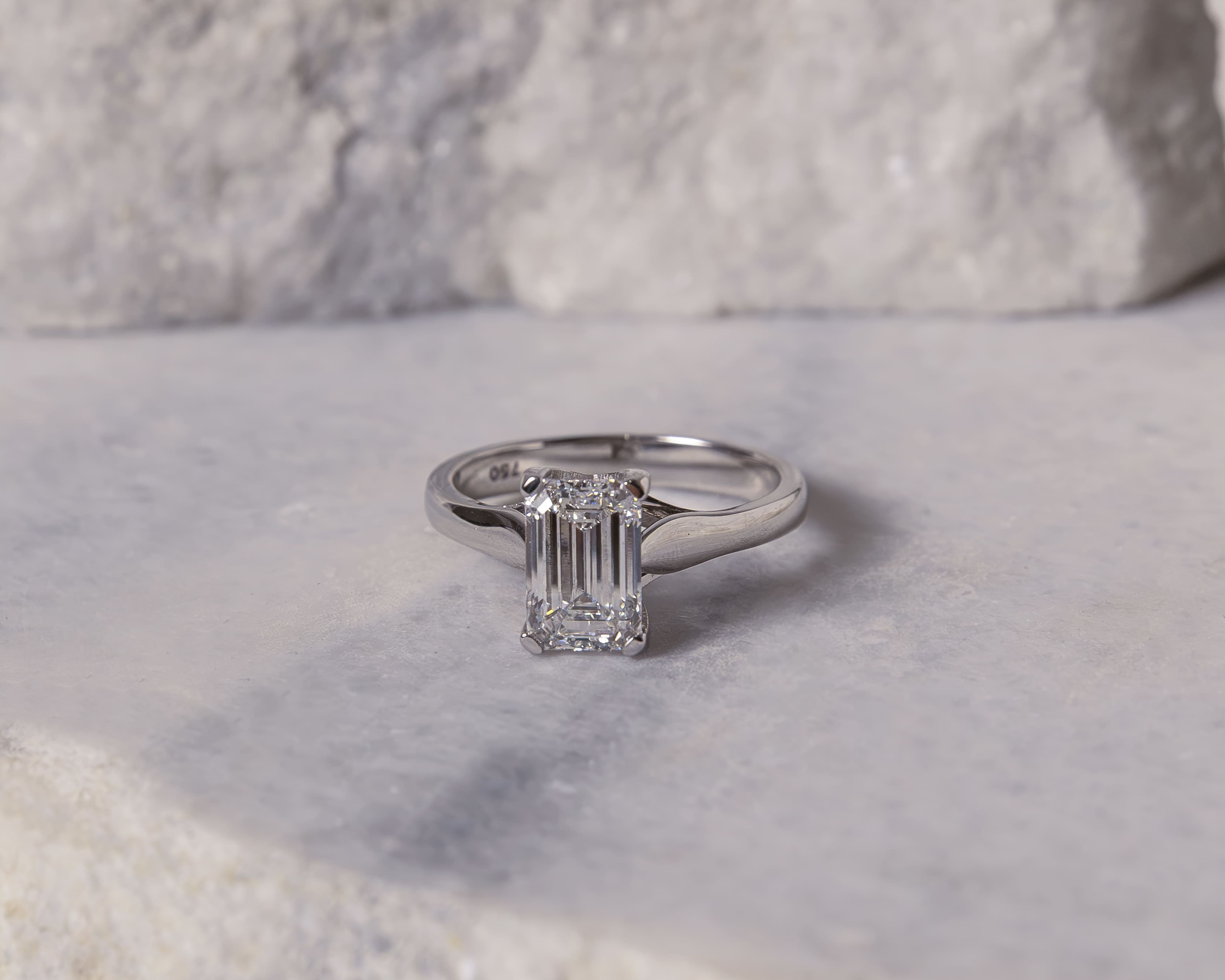 Emerald-cut diamond engagement ring with a polished silver band resting on a light grey textured surface.