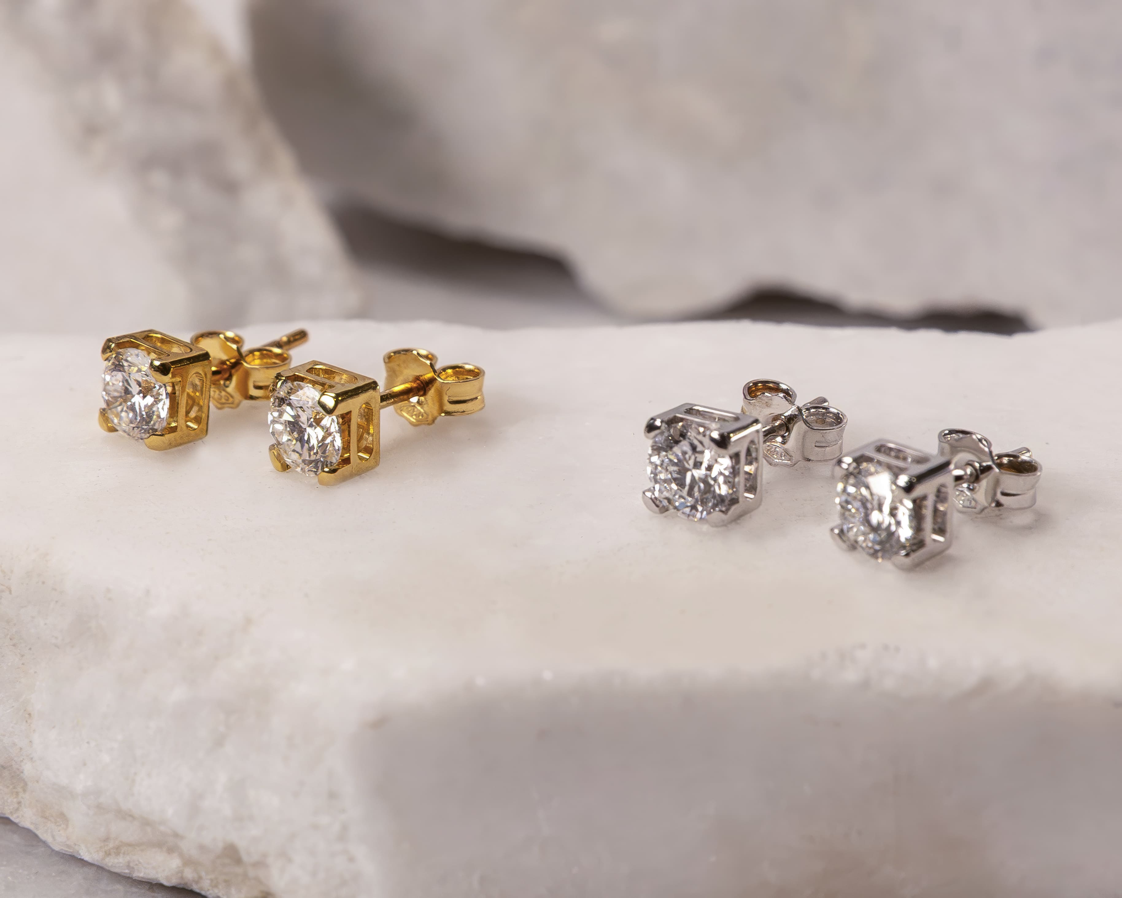 Two pairs of stud earrings with clear round gemstones, one set in gold and the other in silver, displayed on a white stone surface.