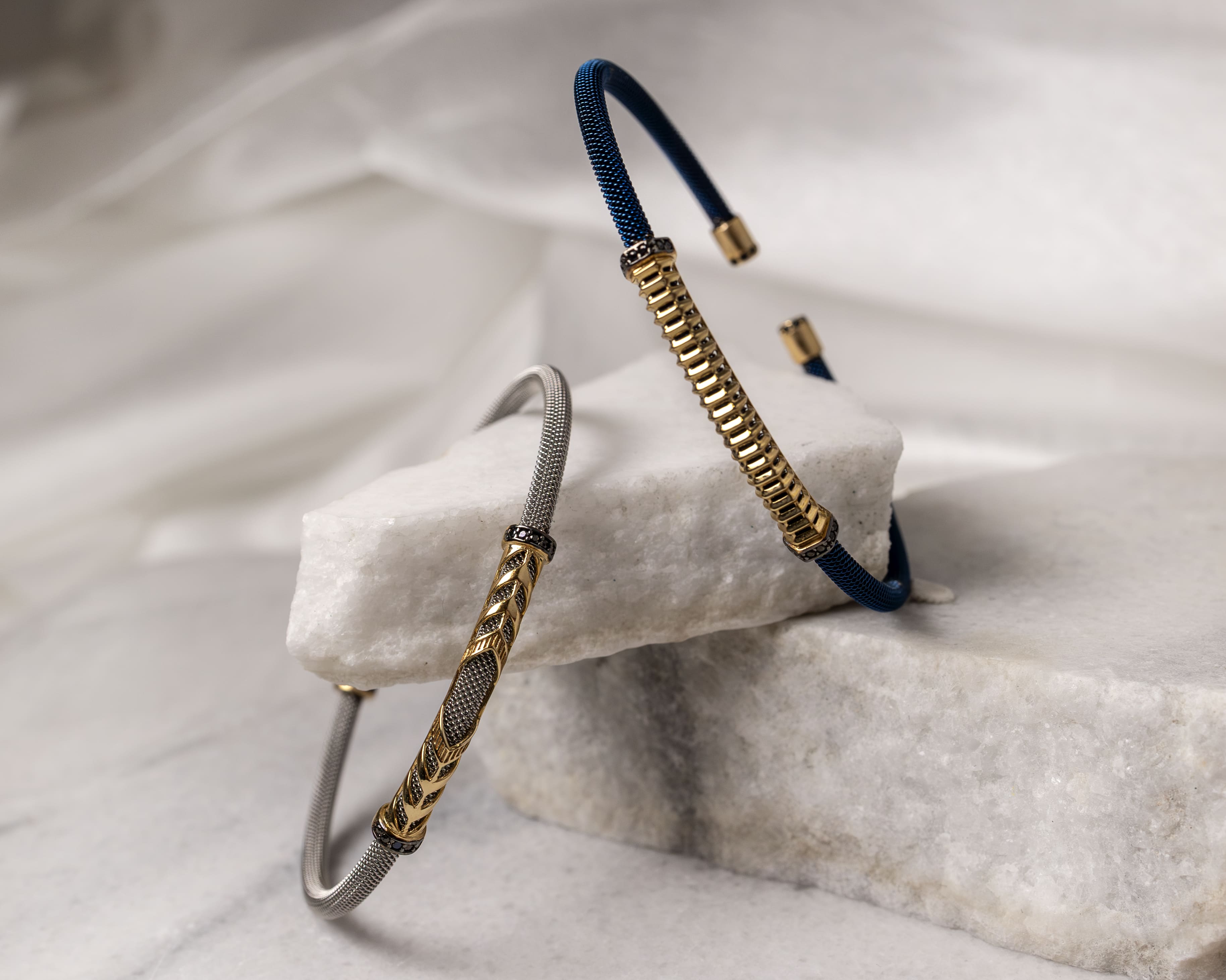 Two elegant bracelets with gold and mesh designs displayed on white marble blocks.